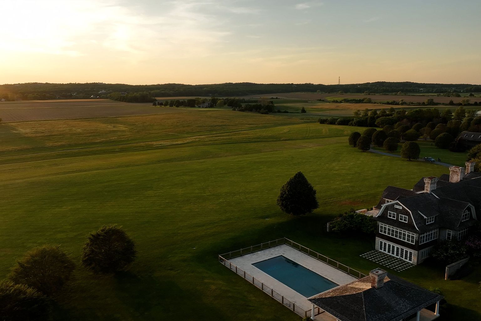 This aerial shot showcases a sprawling estate with a meticulously maintained lawn, a rectangular swimming pool, and a large, elegant house with a dark roof and white-trimmed windows. The property is surrounded by fields and trees, creating a sense of privacy and seclusion. The warm sunset lighting enhances the overall appeal and highlights the property's grandeur.