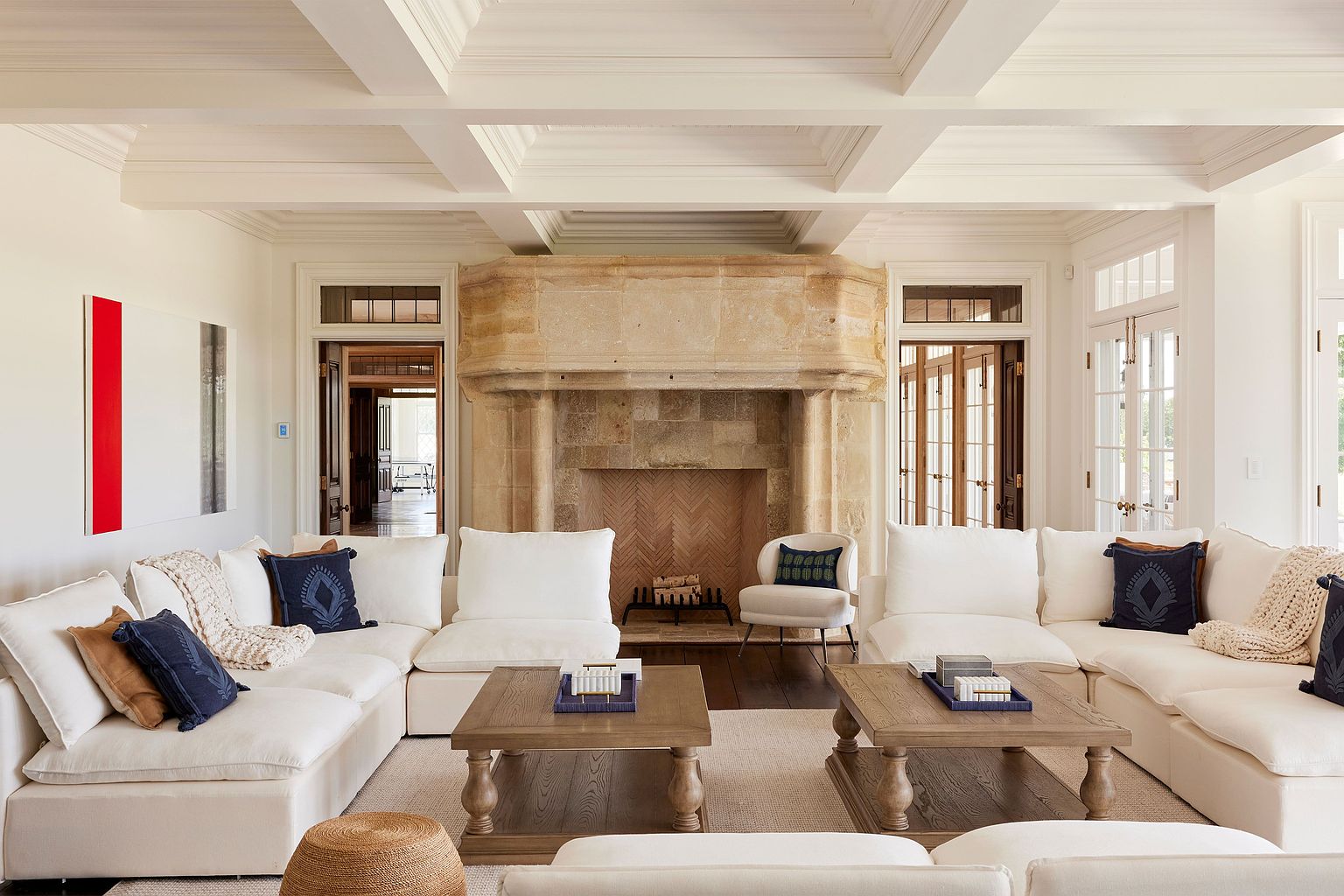 This is a spacious and luxurious living room featuring a large, comfortable white sectional sofa arranged around two wooden coffee tables. A grand stone fireplace serves as a focal point, flanked by doorways leading to other rooms. The room is well-lit with natural light and features elegant architectural details such as coffered ceilings, creating a sophisticated and inviting atmosphere.