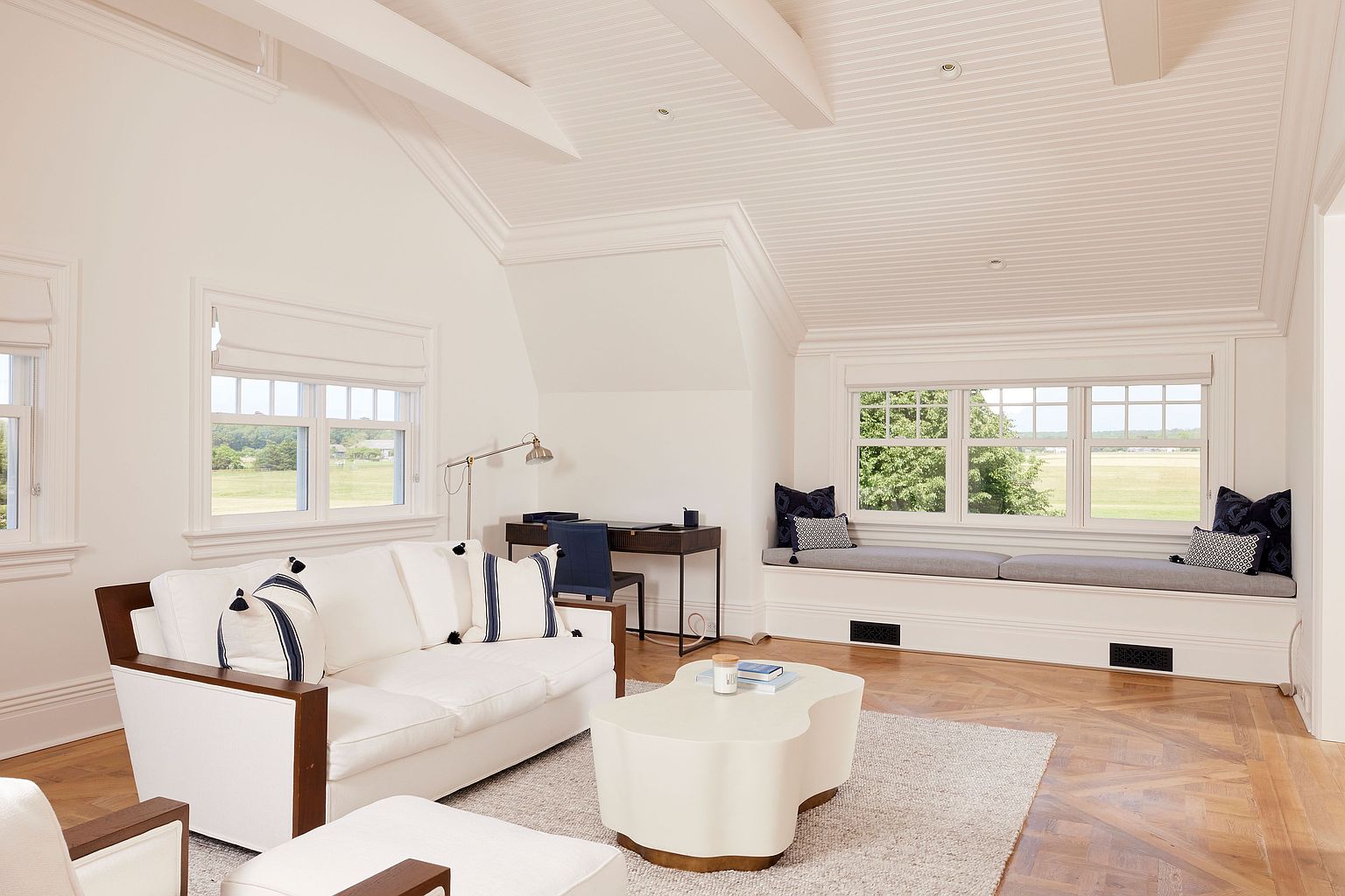 This is an interior shot of a bright and airy living room featuring a white sofa with navy accents, a uniquely shaped coffee table, and a built-in window seat with cushions. The room has a high, angled ceiling with exposed beams and multiple windows that provide ample natural light and views of the outdoors. The hardwood floors and neutral color palette create a sophisticated and inviting atmosphere.