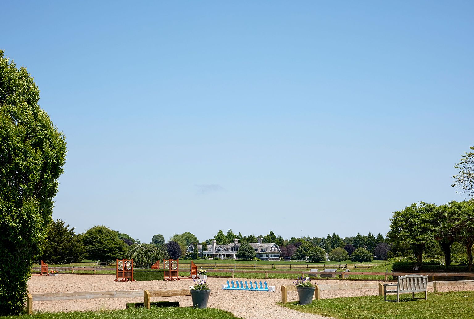 This image showcases a well-maintained yard or garden area, possibly part of a larger estate. The scene includes a sand area with jumps, benches, and potted plants, suggesting an equestrian facility. In the background, a large house is visible, surrounded by trees, under a clear blue sky, creating a serene and upscale atmosphere.