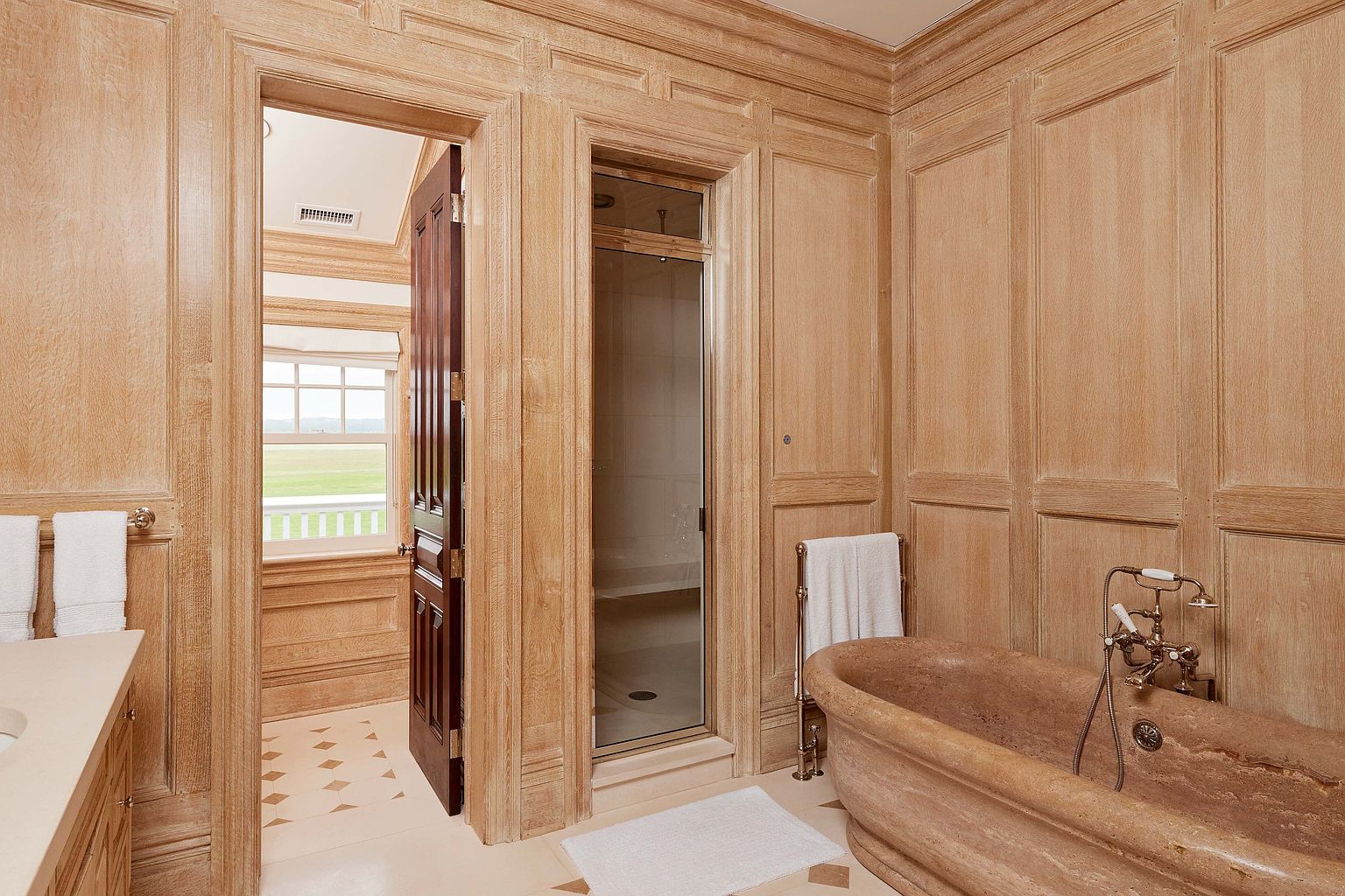 This is a luxurious primary bathroom featuring wood paneled walls, a stone bathtub, and a glass-enclosed shower. The bathroom has a classic and elegant style, with high-end fixtures and finishes. A doorway leads to another room with a view of a green field.