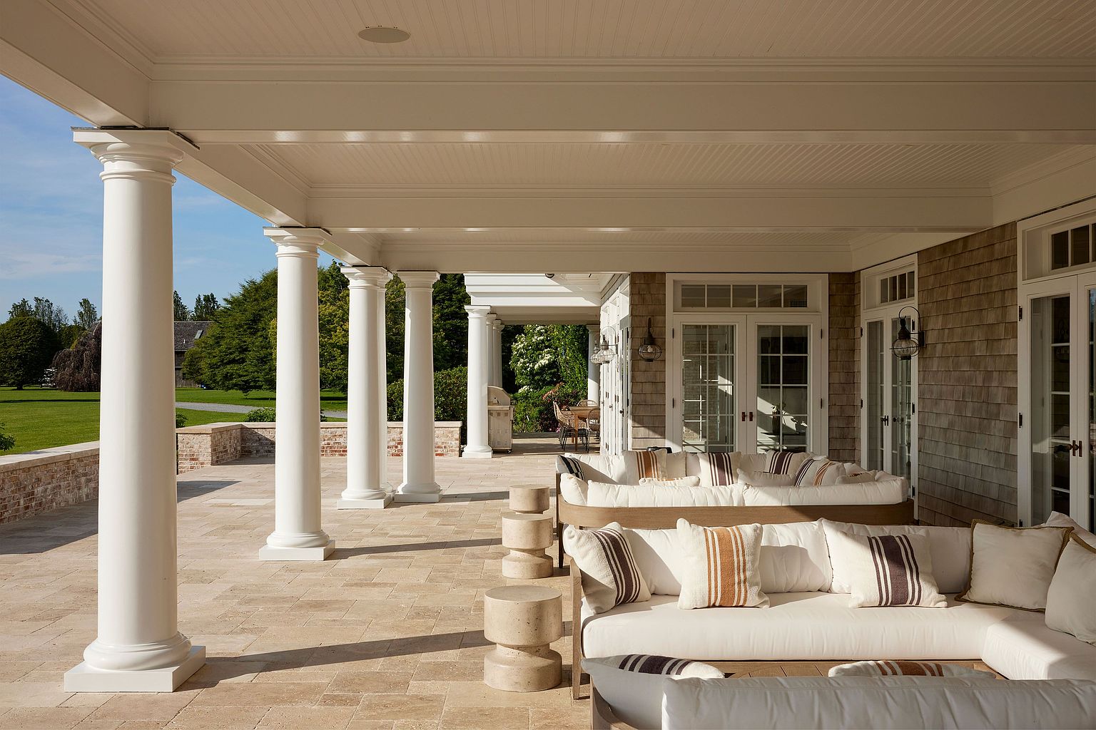 This image showcases a luxurious outdoor patio area, featuring elegant white columns supporting a covered space. The patio is furnished with comfortable, cushioned seating and small, modern side tables. The flooring is made of light-colored stone tiles, and the overall design exudes a sense of sophisticated relaxation and outdoor living.
