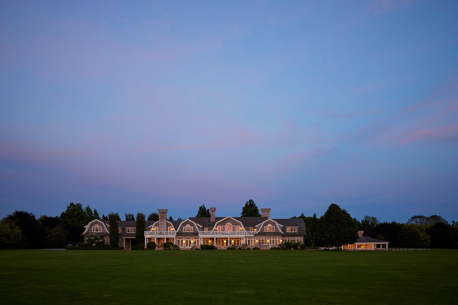 This image showcases the grand front view of a large, luxurious home at dusk. The house features multiple gabled sections, prominent chimneys, and a covered porch that spans a significant portion of the facade. A vast, well-manicured lawn extends from the house, creating a sense of spaciousness and elegance, while the sky transitions from blue to soft pink hues, enhancing the property's serene and upscale ambiance.