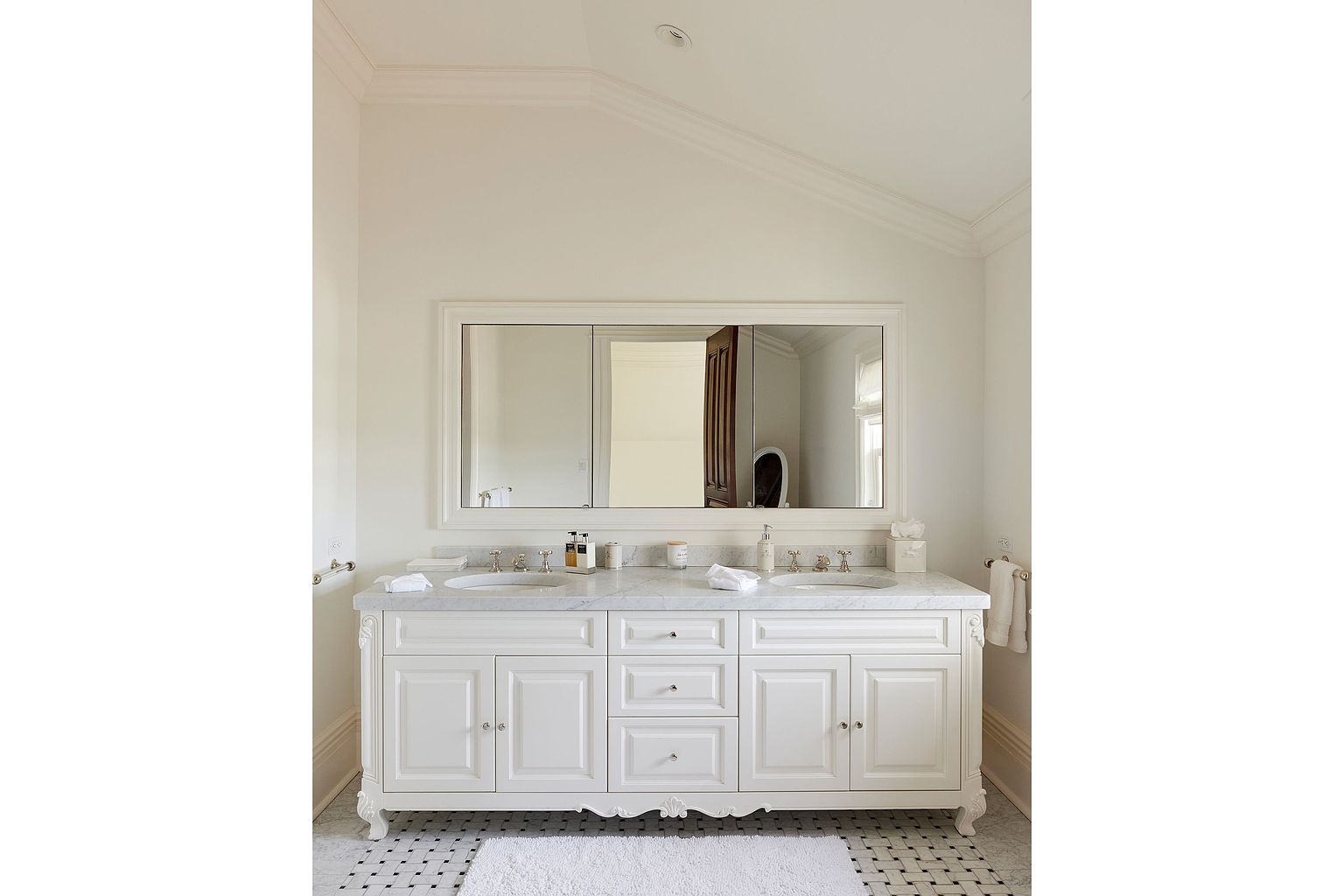 This is a bright and elegant primary bathroom featuring a double vanity with a white marble countertop and classic white cabinetry. A large, framed mirror spans the width of the vanity, reflecting the light and enhancing the spacious feel. The black and white tiled floor adds a touch of sophistication to the room.