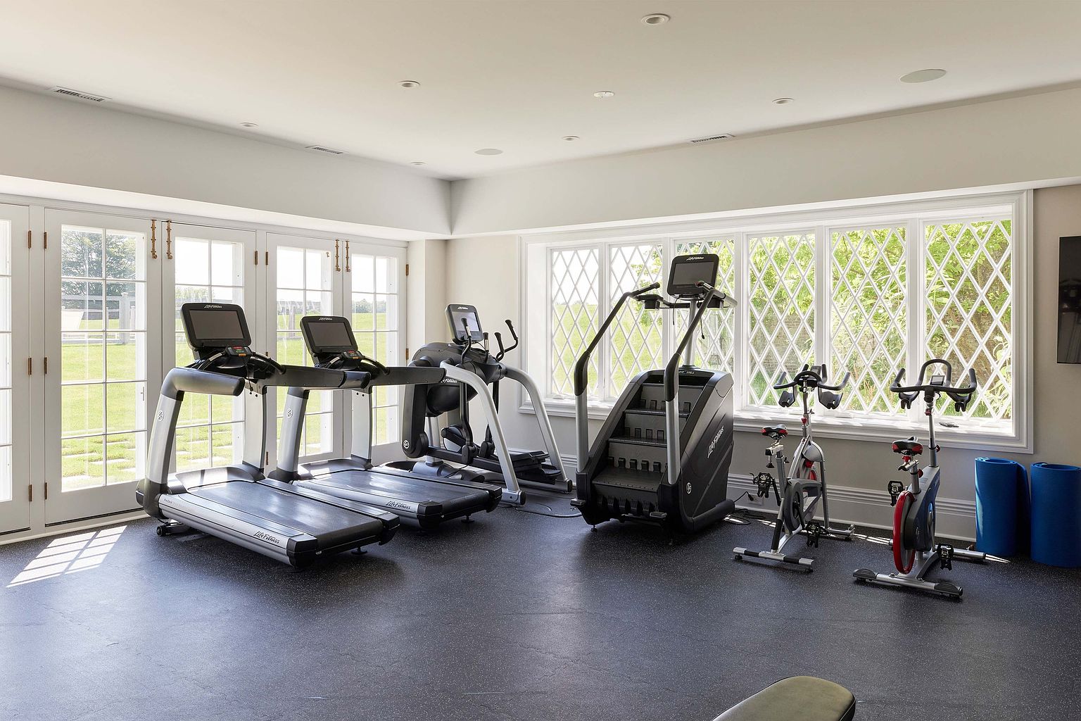 This is a well-equipped home gym featuring multiple treadmills, an elliptical machine, a stair climber, and stationary bikes. Large windows provide ample natural light and a view of the outdoors. The room has a clean and modern aesthetic with a dark rubber floor, creating a dedicated space for fitness.