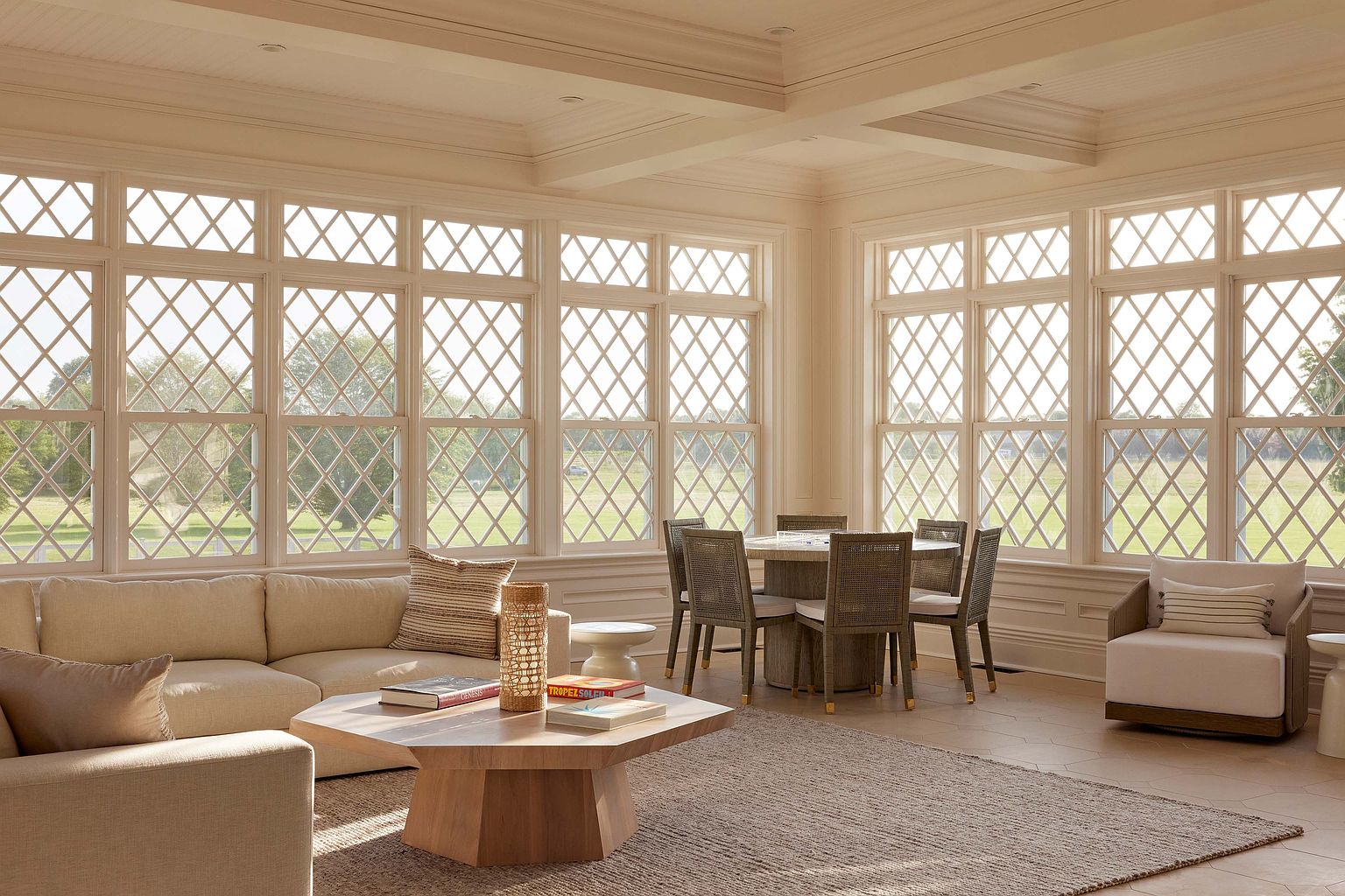This sunroom features large diamond-patterned windows that flood the space with natural light, showcasing a serene outdoor view. The room is furnished with a comfortable beige sofa, a round dining table with woven chairs, and a light-colored area rug, creating a bright and inviting atmosphere. The neutral color palette and classic architectural details enhance the room's elegance and appeal.