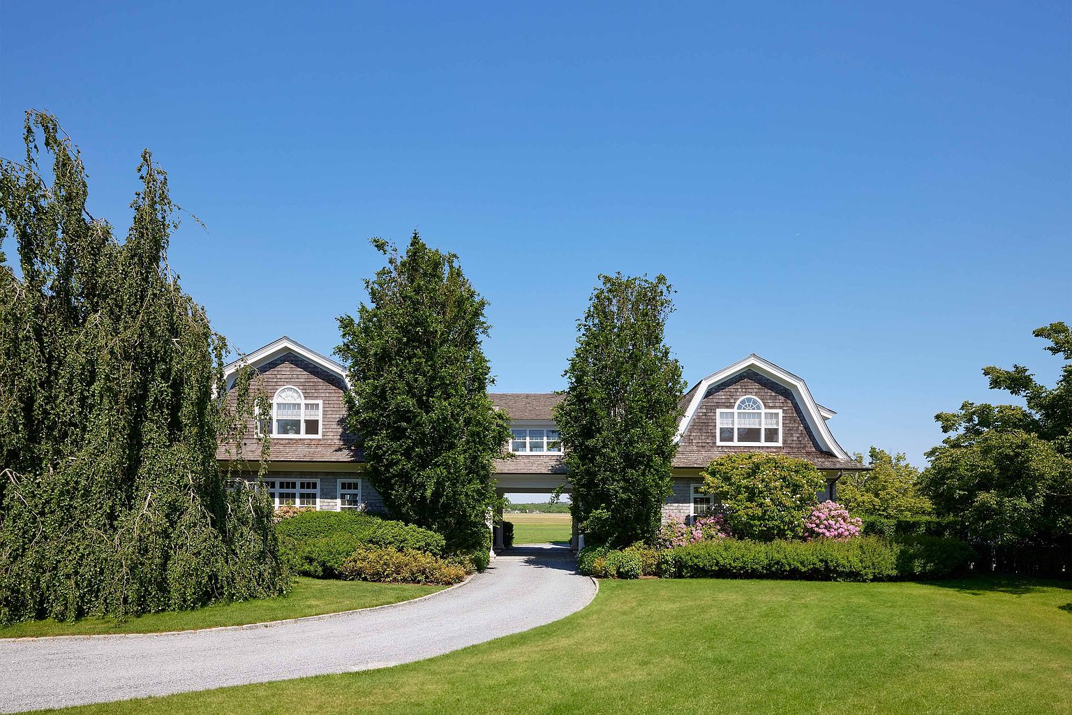 This is a front view of a charming house with a gravel driveway leading up to it. The house features a unique architectural design with dormer windows and a connecting structure between the two main sections. Lush green lawns and mature trees frame the property, creating a welcoming and picturesque setting.
