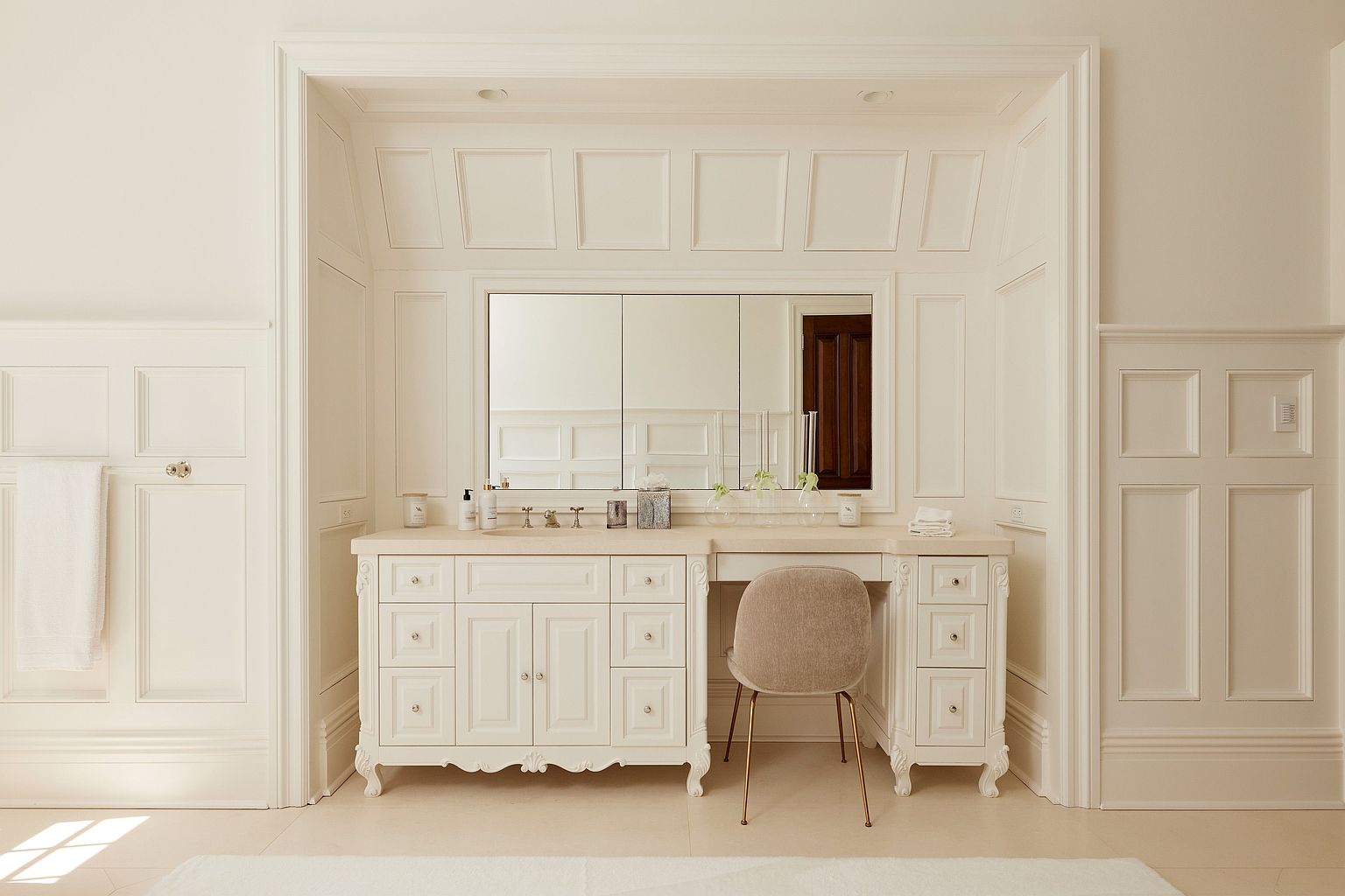 This is a well-lit primary bathroom featuring a vanity with a large mirror and ample storage. The vanity is adorned with decorative knobs and has a classic, elegant design. The walls are paneled, adding to the room's sophisticated aesthetic, and a chair is tucked under the vanity, suggesting a dedicated makeup area.
