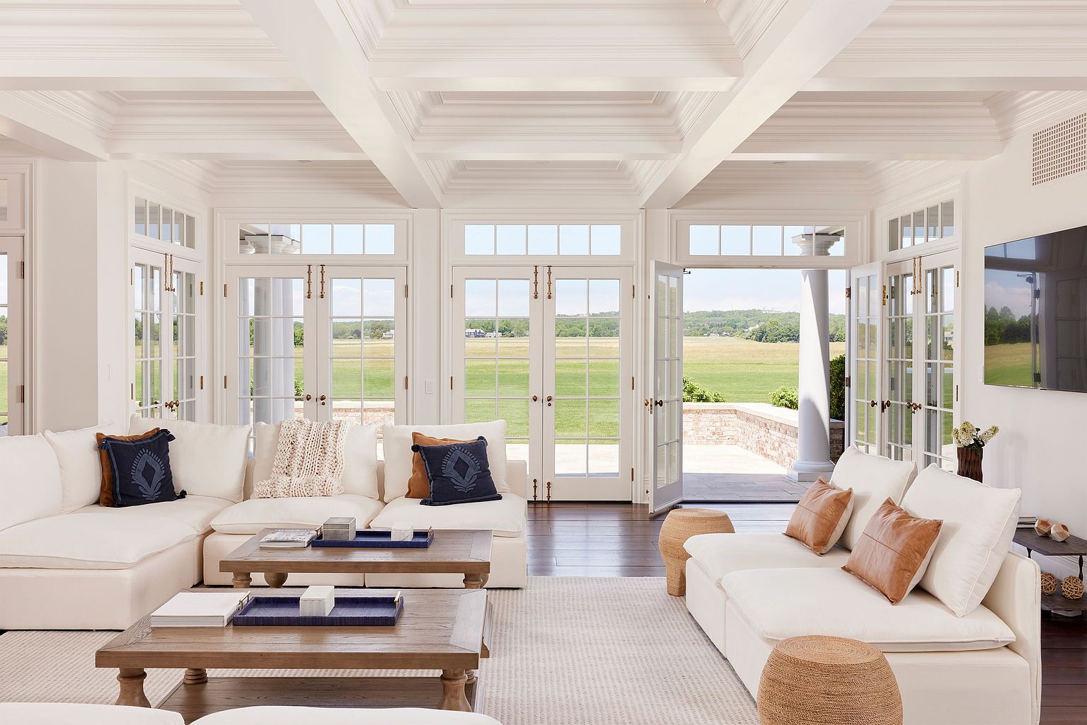 This is a bright and airy living room featuring white sofas, wooden coffee tables, and multiple French doors that open to a green lawn. The room has a sophisticated and comfortable feel, enhanced by the natural light and neutral color palette. The coffered ceiling adds an architectural detail that elevates the space.