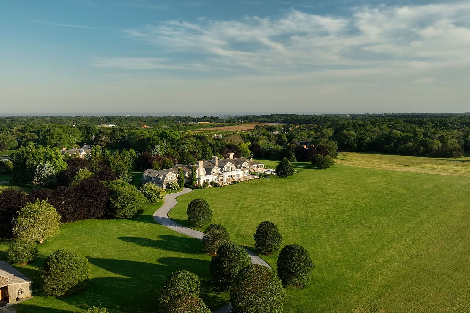 This aerial view showcases a sprawling estate with a grand house, meticulously manicured lawns, and lush surrounding greenery. A winding driveway leads to the residence, emphasizing its secluded and private setting. The overall impression is one of luxury, space, and tranquility, ideal for a discerning buyer seeking a prestigious property.