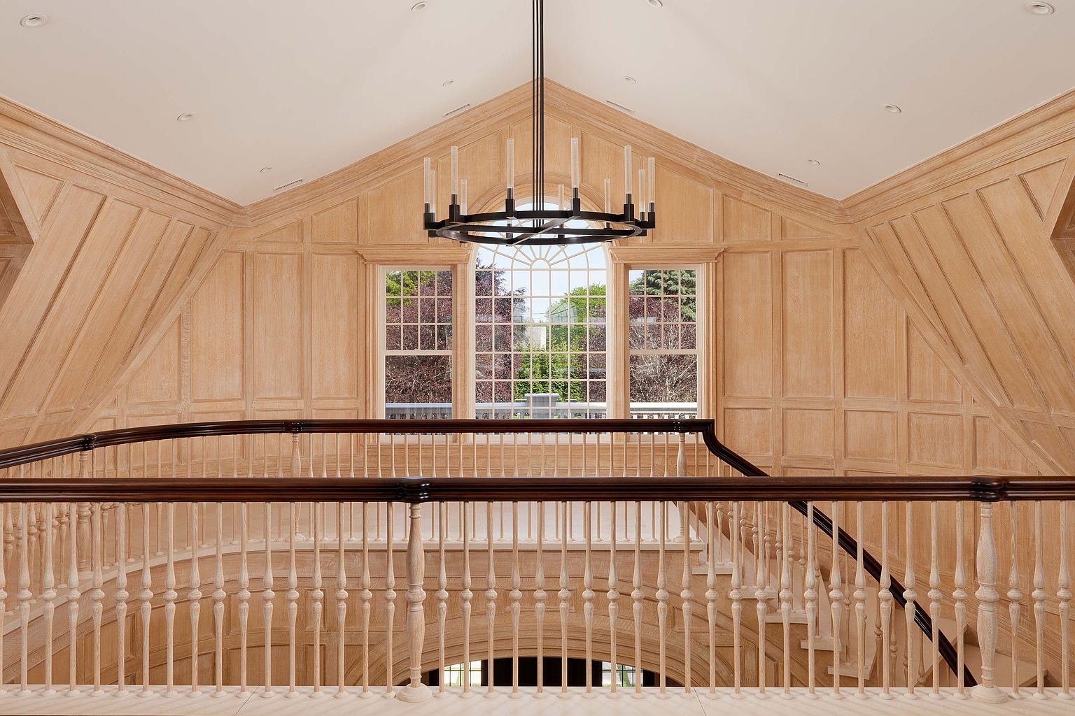 This interior shot showcases a grand hallway and staircase, featuring light wood paneling and a dark wood railing. A modern chandelier hangs above, and large windows provide natural light. The overall impression is one of spaciousness and elegant design.