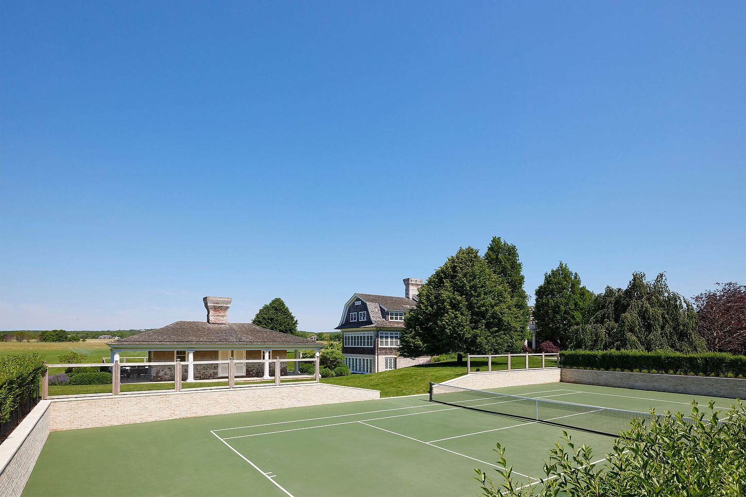 This exterior shot showcases a well-maintained yard featuring a tennis court, manicured hedges, and a charming outbuilding with a stone chimney. A larger house is visible in the background, surrounded by mature trees. The scene exudes a sense of luxury and privacy, perfect for a high-end real estate listing.