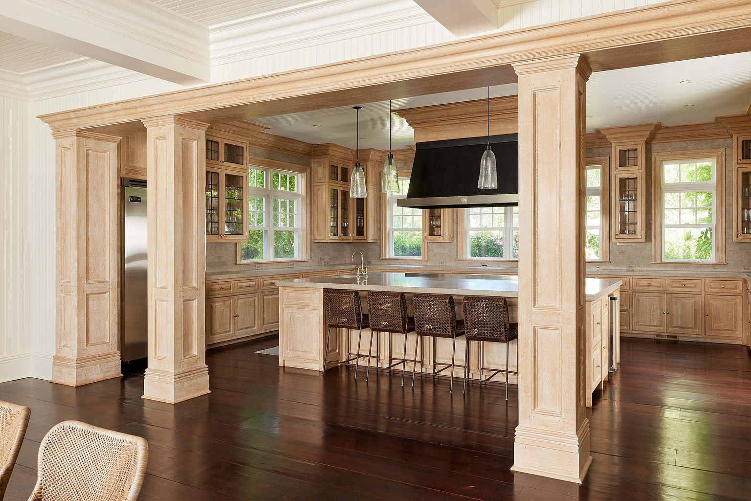 This is a spacious kitchen featuring custom light wood cabinetry, a large central island with seating, and stainless steel appliances. The kitchen is well-lit with natural light from multiple windows and pendant lighting above the island. The dark wood flooring adds a touch of elegance to the space.