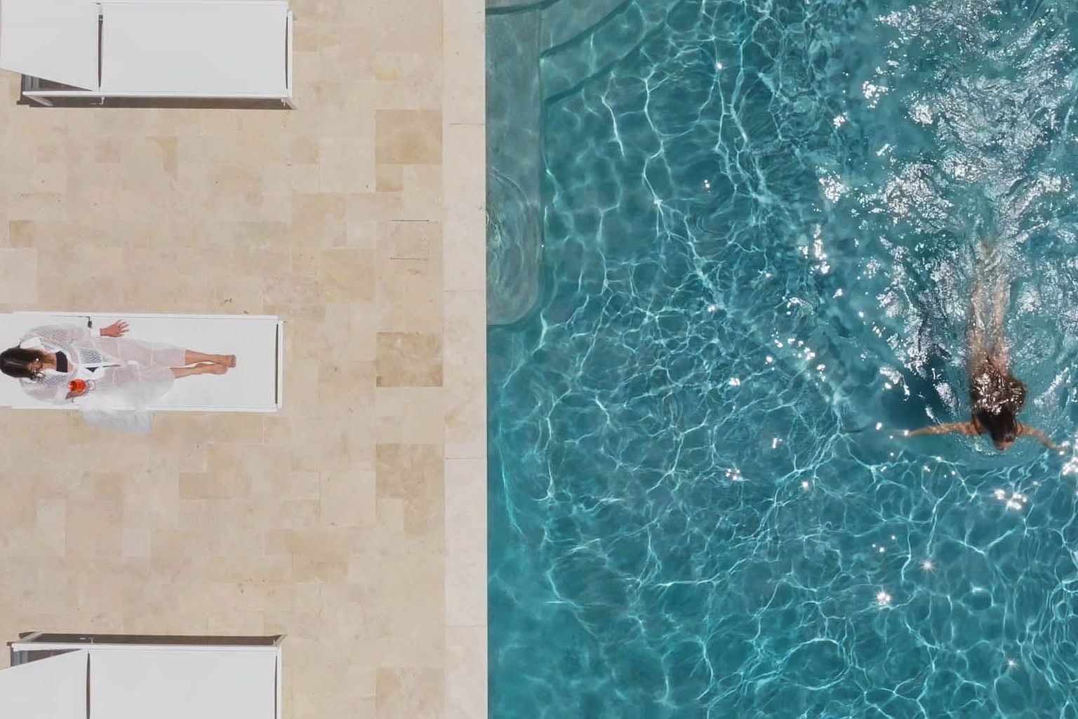 This aerial shot showcases a luxurious pool area with a person swimming in the turquoise water. Adjacent to the pool is a tiled patio area featuring white lounge chairs, one occupied by a person relaxing. The scene evokes a sense of relaxation and upscale leisure, perfect for highlighting the property's amenities.