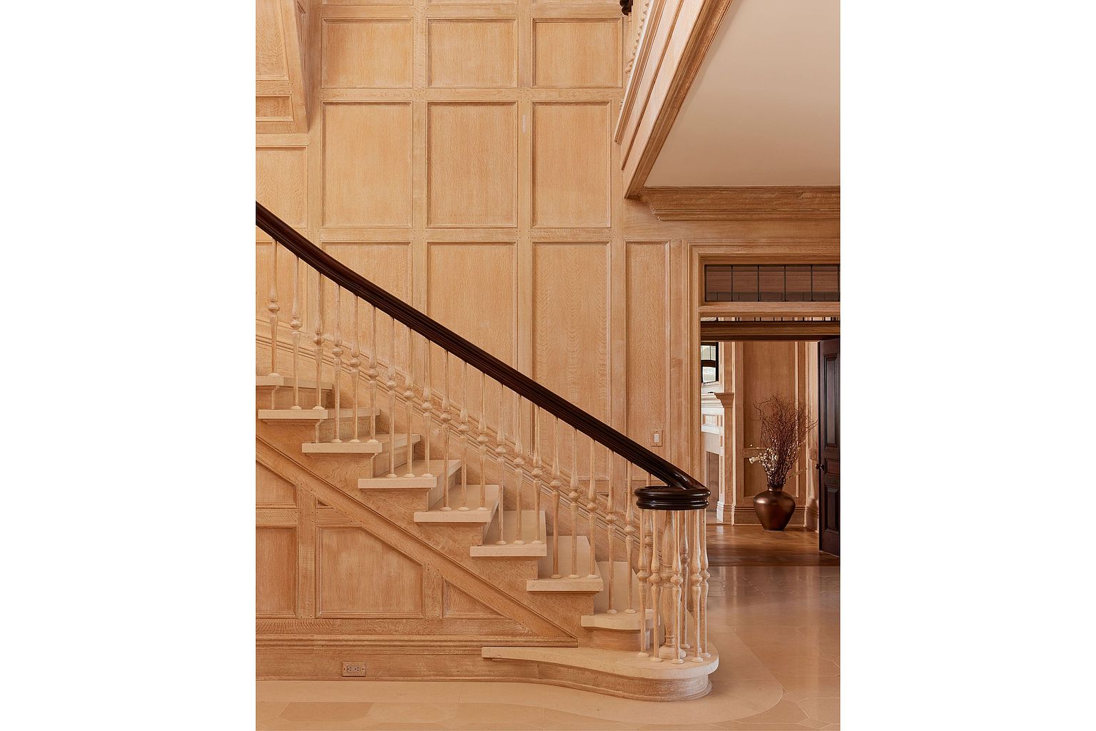 This interior shot showcases a grand hallway with a prominent staircase. The walls are adorned with detailed wood paneling, creating a warm and luxurious atmosphere. The staircase features a dark wood handrail contrasting with the light-colored steps and balusters, leading to an upper level. A glimpse into adjacent rooms reveals hardwood flooring and a sophisticated design aesthetic, suggesting a high-end property.