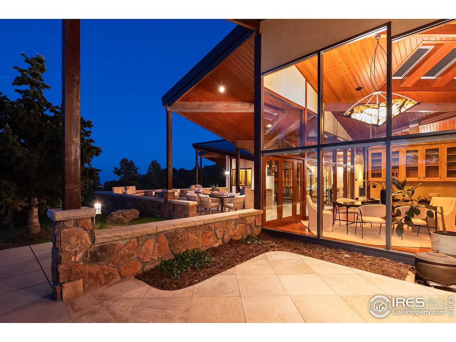 This is an exterior shot of a modern home's patio area at dusk. The patio features a stone retaining wall, a tiled patio surface, and outdoor seating areas. Large windows offer a view into the home's interior, showcasing the open floor plan and wooden ceiling. The overall impression is one of luxury and seamless indoor-outdoor living.