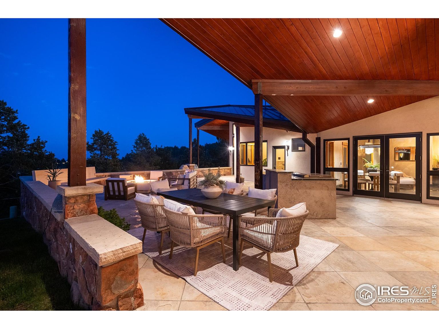 This is a beautifully designed outdoor patio area, perfect for entertaining or relaxing. The space features a dining table with wicker chairs, a cozy fire pit area with comfortable seating, and a built-in outdoor kitchen. The patio is paved with stone tiles and is partially covered by a wooden roof, providing shade and shelter.