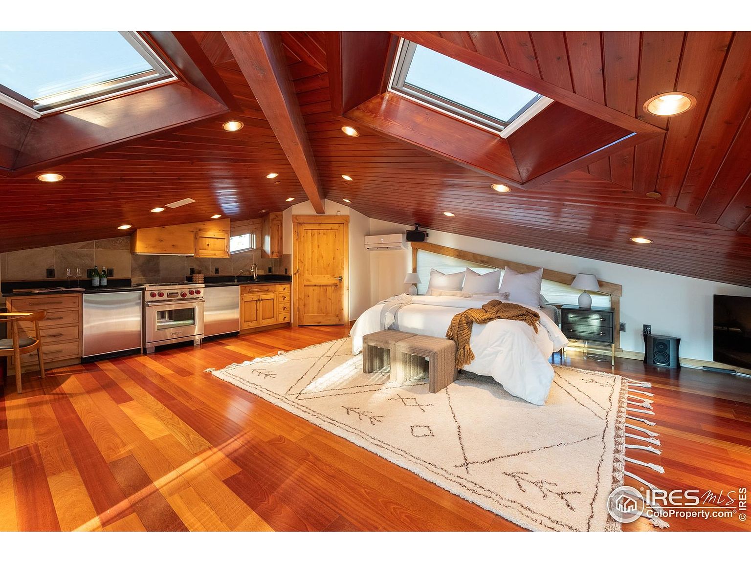 This is an interior shot of a primary bedroom featuring a unique attic-style design with wooden ceilings and beams. The room includes a bed, nightstands, a rug, and a small kitchenette area with stainless steel appliances. Skylights provide natural light, enhancing the warm and inviting atmosphere.
