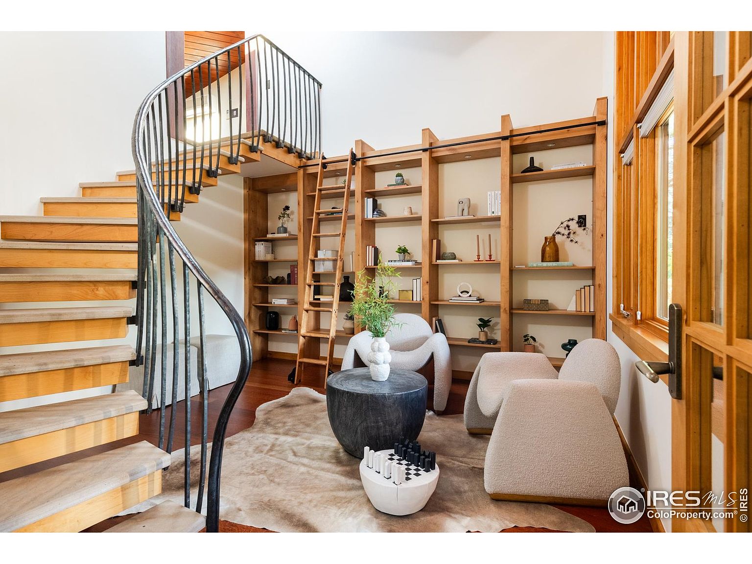 This interior shot showcases a stylish living room with a unique spiral staircase and custom built-in bookshelves. The room features modern furniture, including rounded chairs and a distinctive coffee table, set on a patterned rug. The natural wood tones of the staircase and bookshelves create a warm and inviting atmosphere, highlighting the home's architectural details.