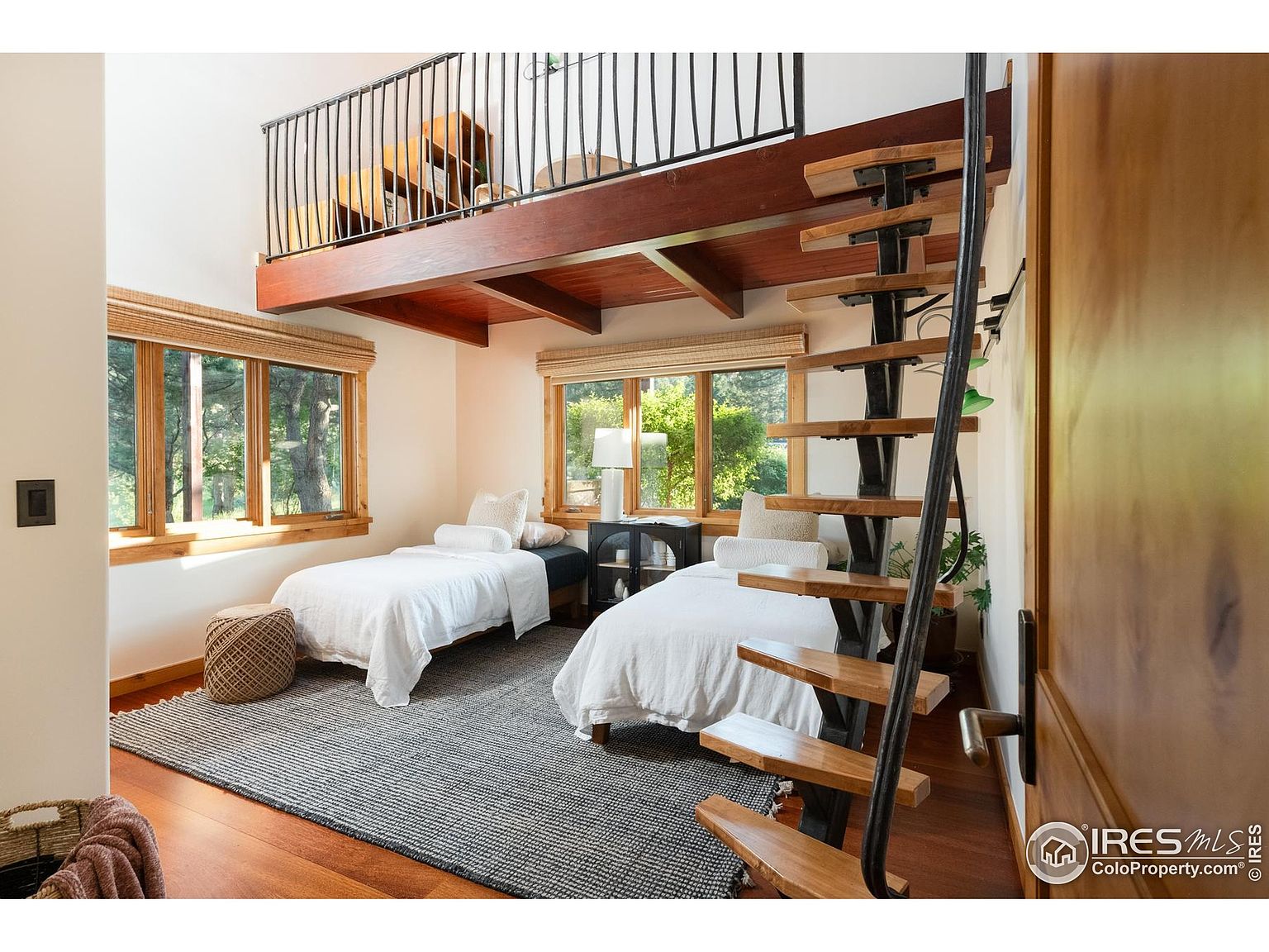 This is a bright and airy guest bedroom featuring two twin beds with white linens, a gray patterned rug, and natural light streaming through the windows. A unique spiral staircase leads to a loft area above, adding architectural interest. The room has a cozy and inviting atmosphere, perfect for guests.