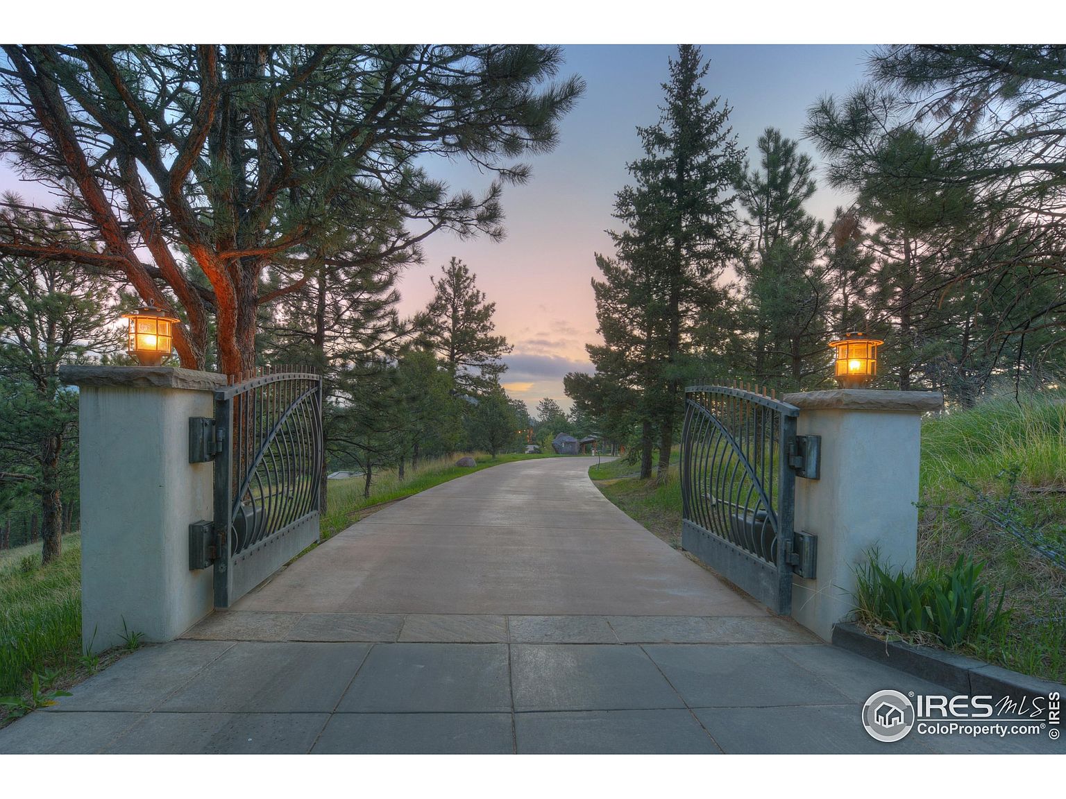 The image showcases an inviting entryway to a property, featuring an open wrought iron gate flanked by stone pillars with illuminated lanterns. A paved driveway stretches into the distance, lined with mature trees and landscaping. The scene evokes a sense of privacy and elegance, suggesting a well-maintained and desirable estate.