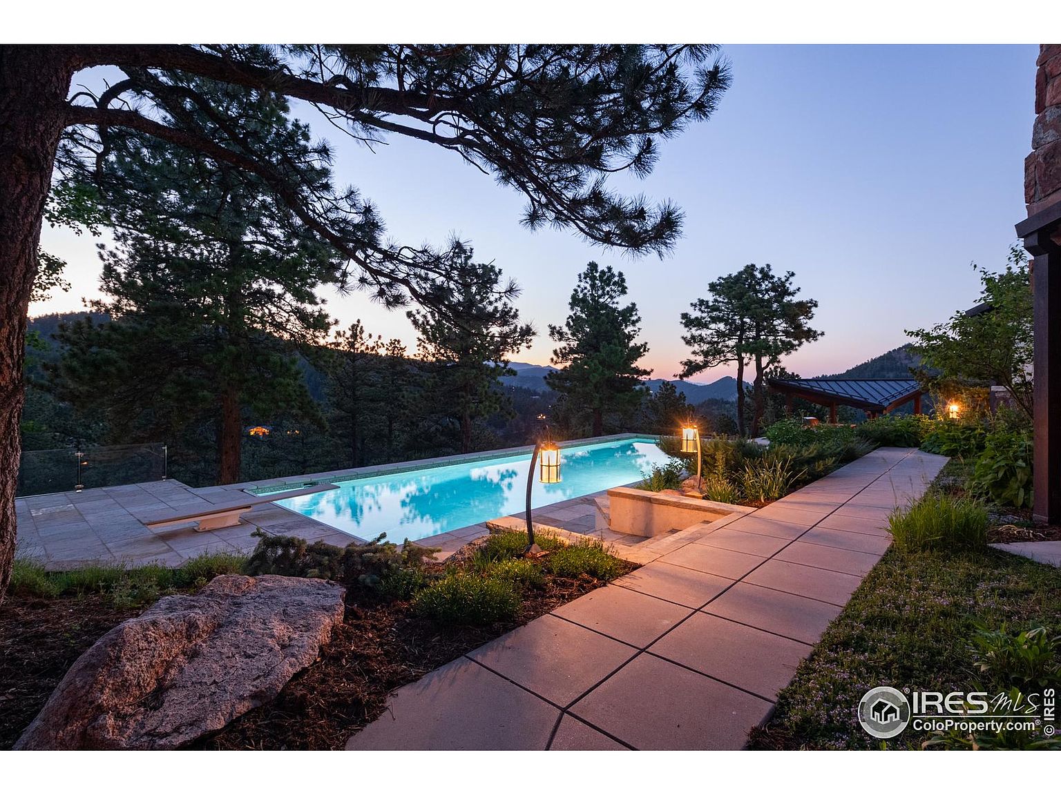 This image showcases a luxurious outdoor pool area at dusk. The pool is surrounded by lush landscaping, including trees and manicured gardens, creating a serene and private setting. A paved walkway leads to the pool, and stylish lanterns provide ambient lighting, enhancing the overall elegance and appeal of the property.