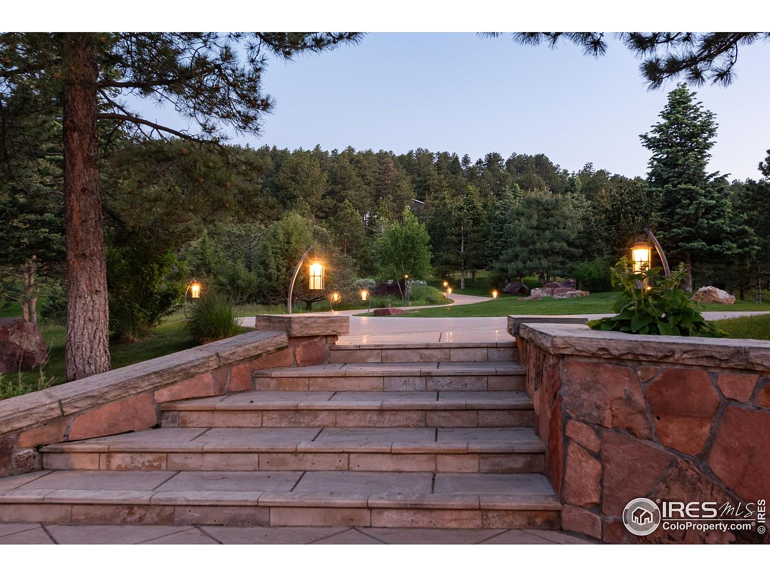 This image showcases a beautifully landscaped yard and garden, featuring stone steps leading up to a pathway illuminated by elegant outdoor lighting. The lush greenery, mature trees, and well-maintained lawn create a serene and inviting atmosphere. The stone retaining walls add a touch of rustic charm and enhance the overall aesthetic appeal of the property.
