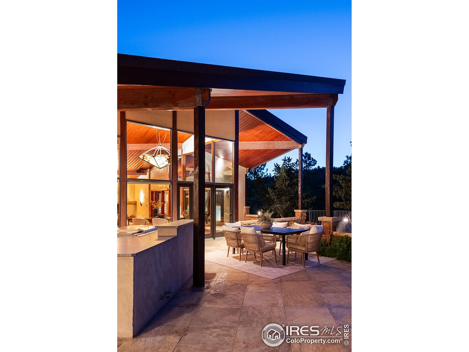 This image showcases a beautifully designed outdoor patio area, featuring a dining set with woven chairs and a dark-topped table, set upon a light-colored rug. The patio is constructed with large stone tiles and is partially covered by a modern, angular roof supported by wooden beams and columns. The space offers a seamless transition between indoor and outdoor living, with large glass windows providing views into the home.