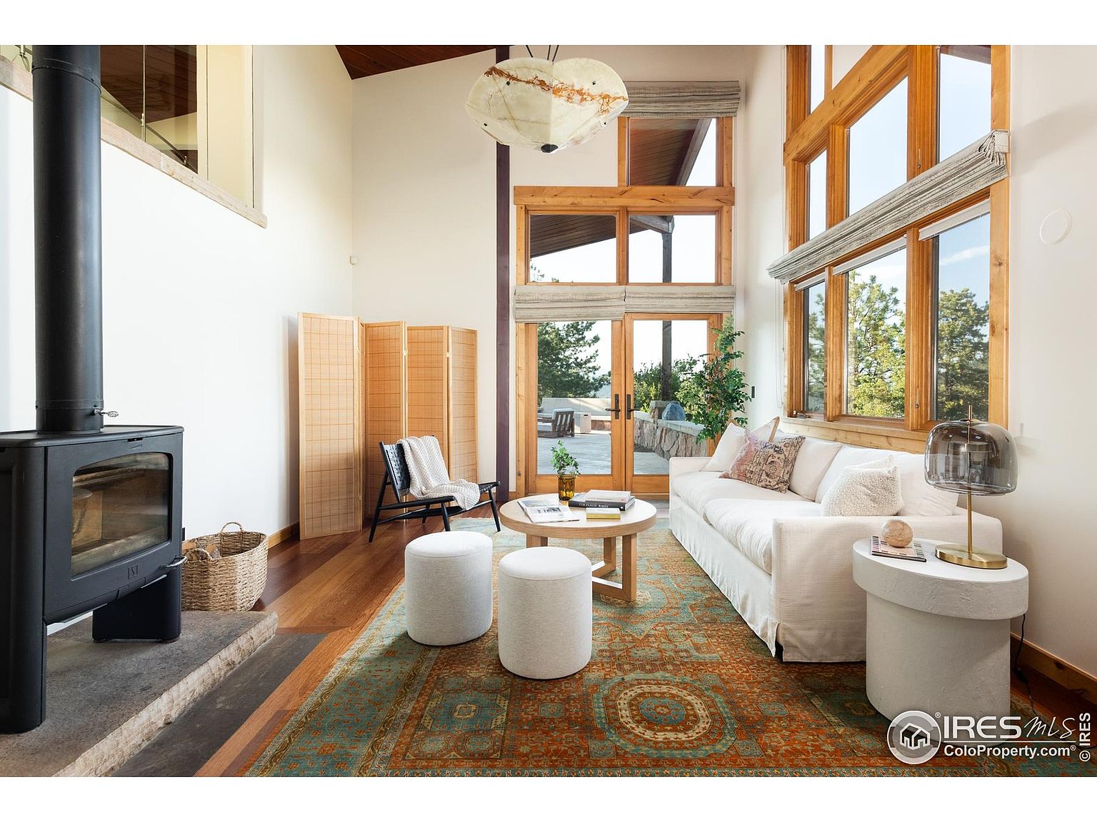 This is an interior shot of a living room featuring a modern fireplace, a white sofa, and large windows that provide ample natural light. The room is decorated with a patterned rug, round ottomans, and a wooden screen, creating a cozy and inviting atmosphere. The high ceilings and wooden window frames add a touch of rustic charm to the contemporary design.