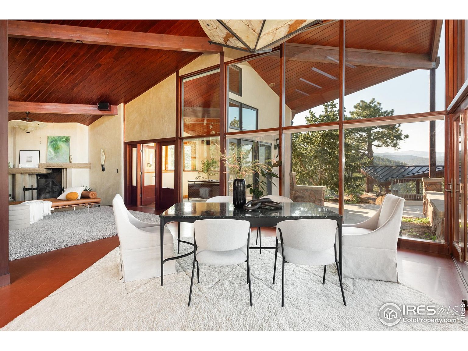 This is an interior shot of a dining room featuring a dark marble table with modern white chairs. The room has floor-to-ceiling windows offering a view of the outdoors, and the ceiling is wood paneled with exposed beams. A large white rug covers the floor, adding to the room's bright and airy feel.