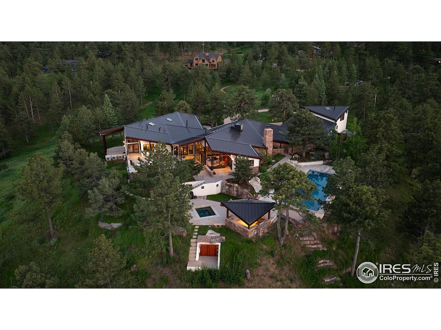 This aerial shot showcases a luxurious estate nestled among lush greenery. The property features a modern architectural design with a dark roof, complemented by a swimming pool and spa area. The surrounding landscape provides privacy and a sense of seclusion, making it an ideal retreat.