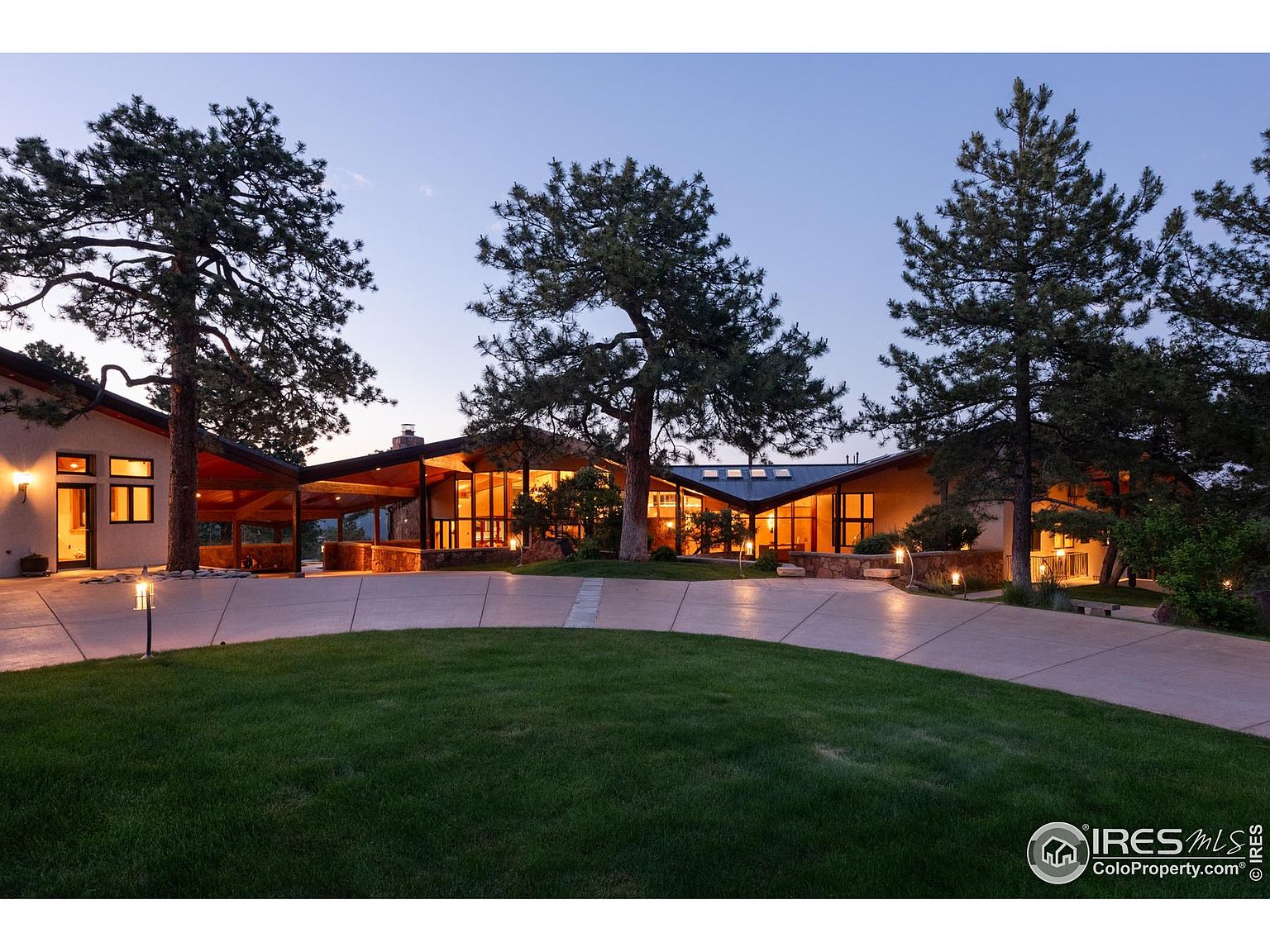 This is a front exterior view of a sprawling, modern home at dusk. The house features a unique roofline, large windows that glow with interior light, and a well-manicured lawn. Mature trees frame the property, adding to its privacy and curb appeal, while a long driveway leads to the entrance.