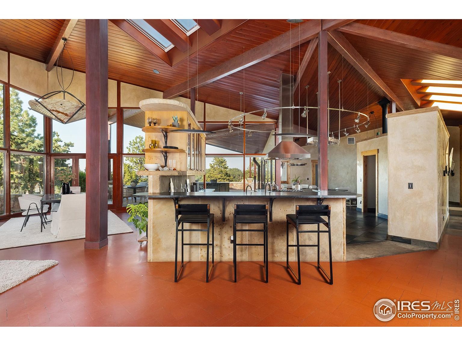 This interior shot showcases a unique kitchen with a striking architectural design, featuring exposed wooden beams and a high ceiling with skylights. The kitchen island, complete with bar stools, anchors the space, while large windows offer expansive views of the outdoors. The warm-toned flooring and natural materials create an inviting and sophisticated atmosphere.
