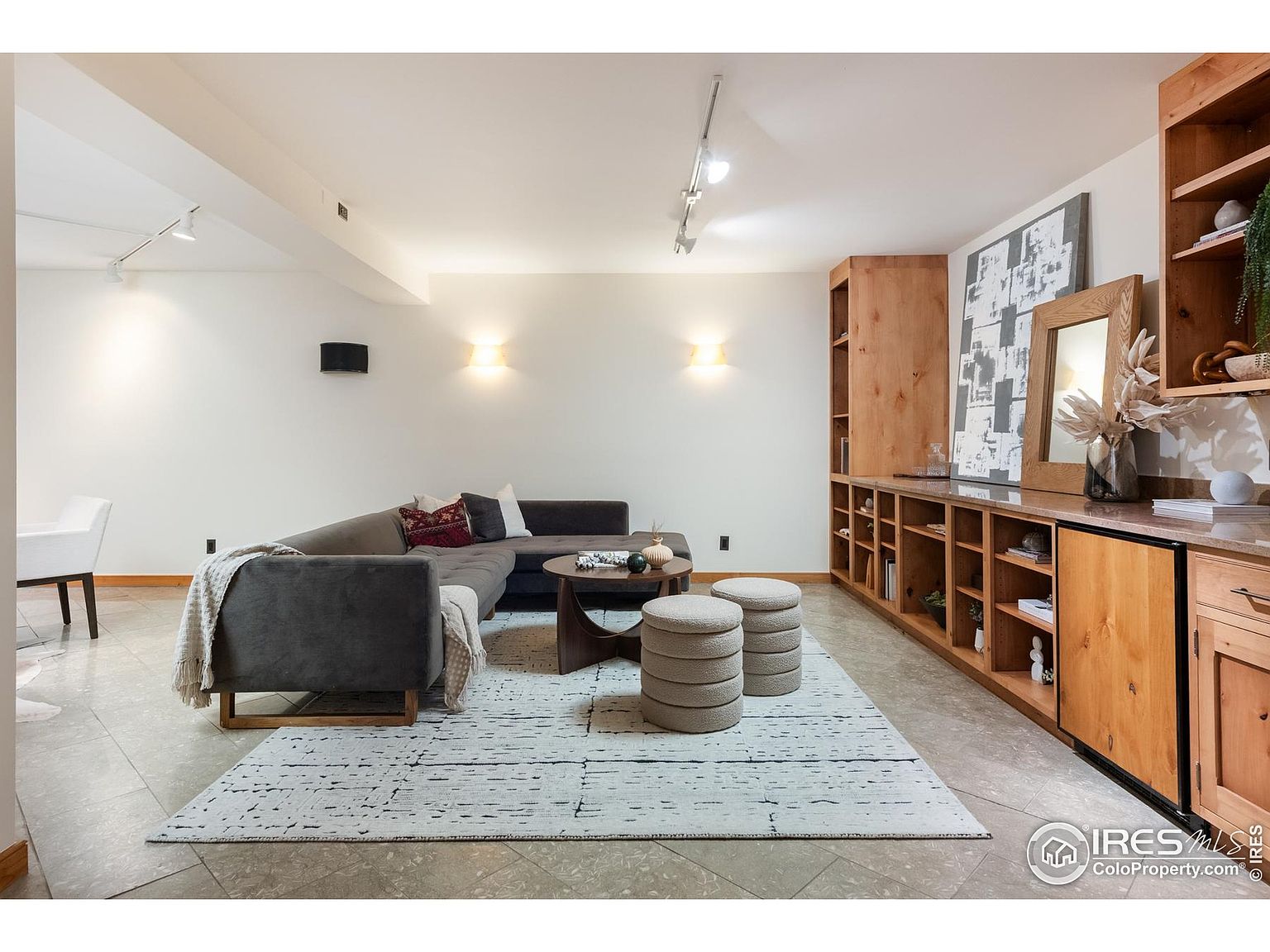 This interior shot showcases a stylish living room with a comfortable sectional sofa, a round coffee table, and two stacked ottomans on a patterned rug. Built-in wooden shelving and cabinets line one wall, providing ample storage and display space. The room is well-lit with a combination of recessed lighting and wall sconces, creating a warm and inviting atmosphere.