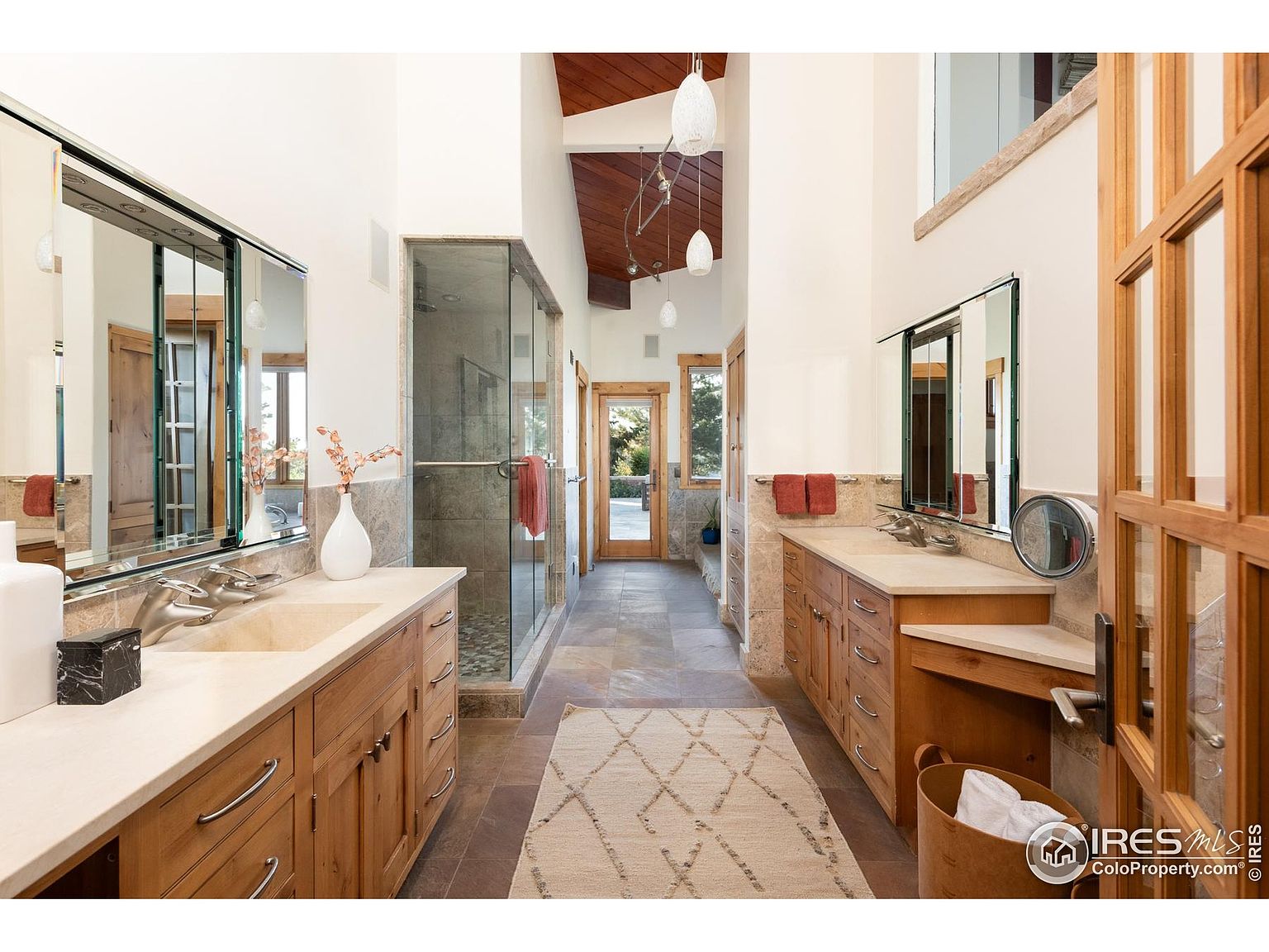This is a well-lit primary bathroom featuring dual vanities with light wood cabinetry and beige countertops. A glass-enclosed shower is visible in the background, and the space is decorated with neutral tones and a patterned rug. The bathroom has a luxurious and spacious feel, with a view to the outside.