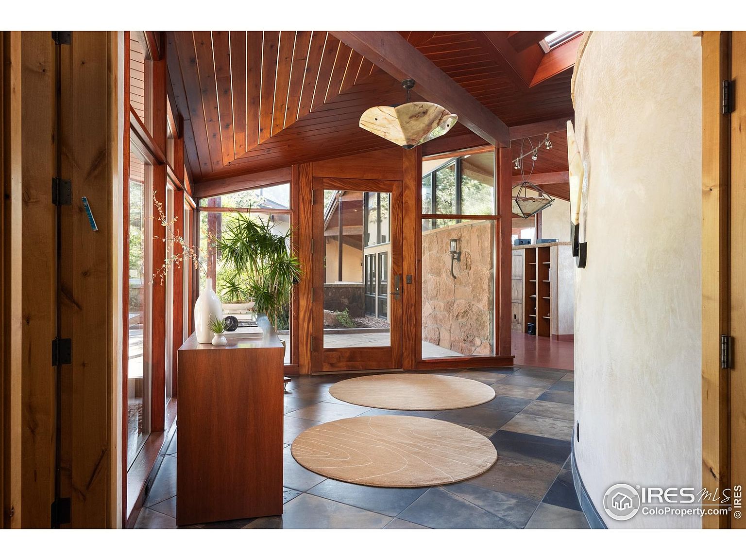This interior shot showcases a unique hallway with a blend of natural wood and stone elements. The space features a high, angled wooden ceiling, large windows, and a curved wall, creating an open and airy feel. Two round rugs soften the tiled floor, adding warmth and texture to the area.