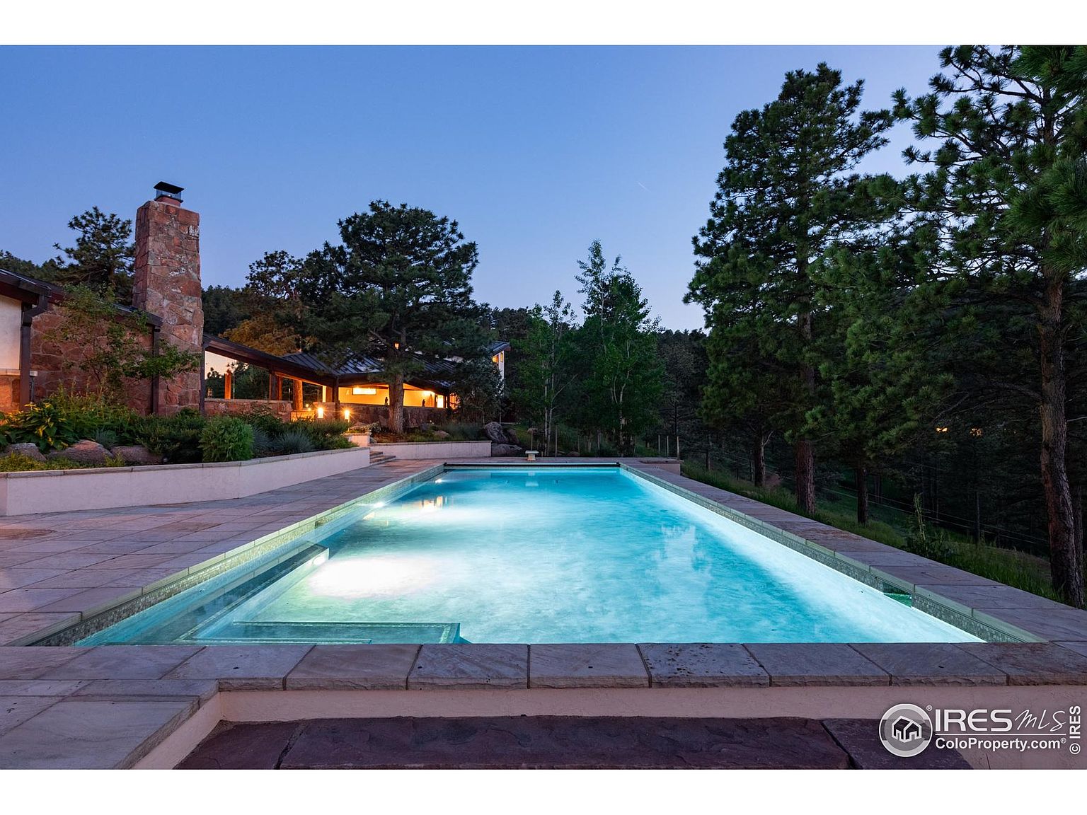 This image showcases a luxurious outdoor pool area at dusk. The rectangular pool is illuminated, creating a serene and inviting atmosphere. The surrounding stone patio and lush landscaping enhance the property's appeal, suggesting a private and upscale retreat.