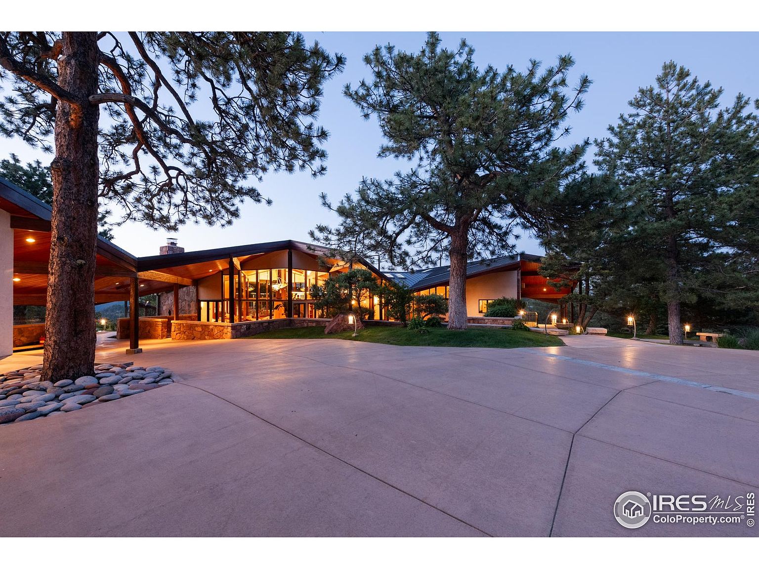 This is a front exterior view of a modern home with a unique architectural design. The house features a low-slung roofline, large windows, and a stone facade. The property is surrounded by mature trees and landscaping, and a spacious driveway leads to the entrance. The lighting suggests it is dusk or dawn, creating a warm and inviting atmosphere.