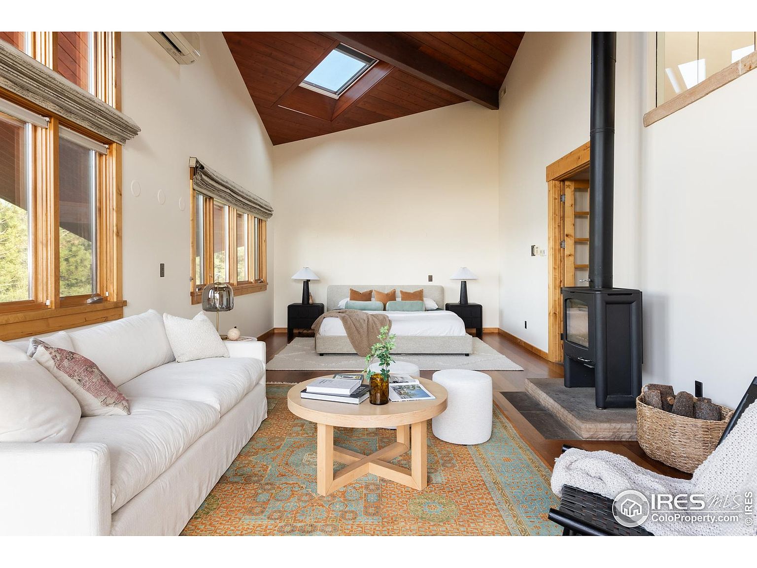 This is a spacious primary bedroom featuring a high vaulted ceiling with a skylight and a modern wood-burning stove. The room is furnished with a light-colored bed, matching nightstands, and a comfortable white sofa. A round coffee table sits on an area rug, adding a touch of warmth and style to the space.