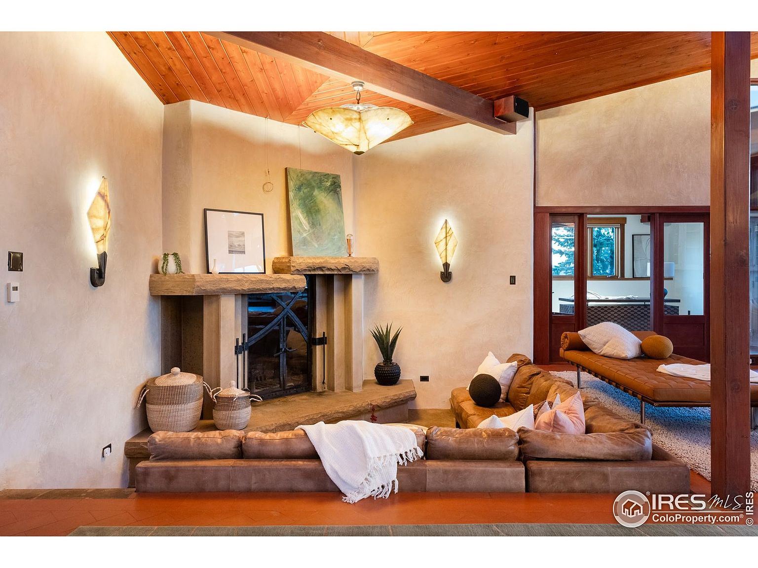 This is an interior shot of a living room featuring a stone fireplace, a brown leather sectional sofa, and a daybed. The room has a warm and inviting atmosphere, enhanced by the wooden ceiling and beams. The lighting fixtures add a unique touch to the space.