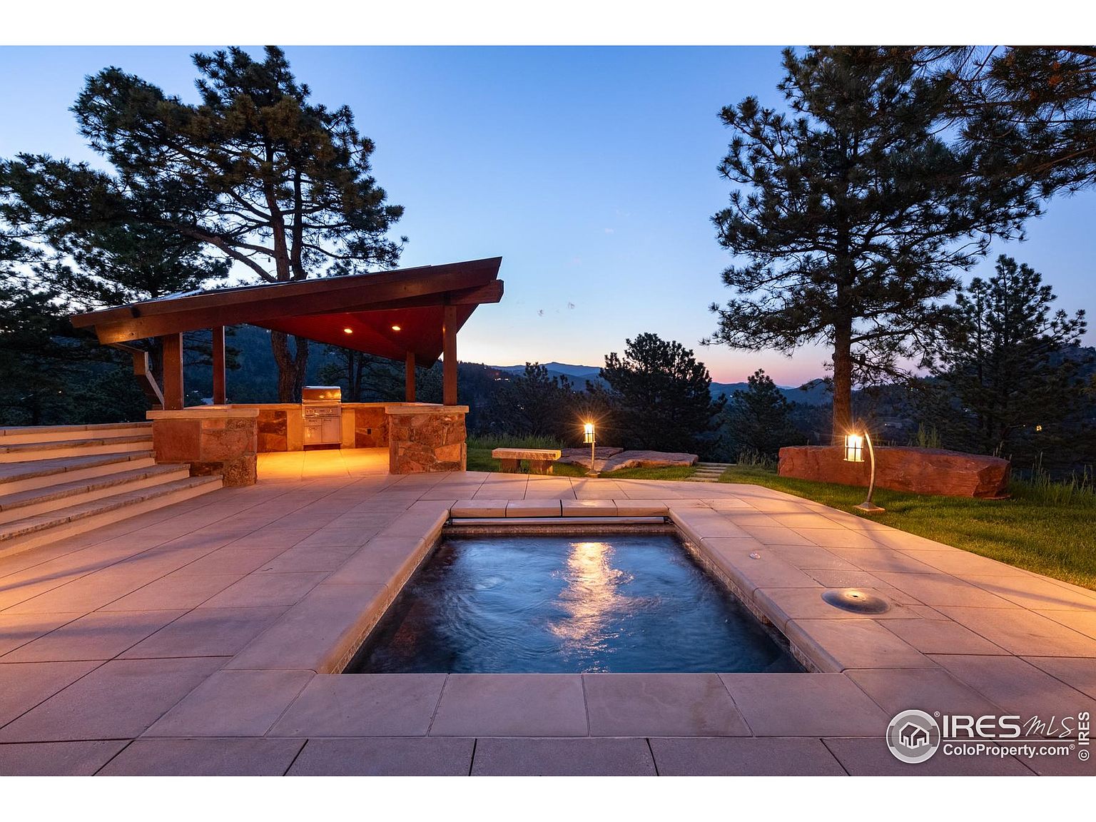 This image showcases a luxurious outdoor pool and spa area at dusk. The rectangular pool is surrounded by stone tiling, and a covered outdoor kitchen with a built-in grill is visible in the background. Landscape lighting illuminates the area, highlighting the trees and creating a serene and inviting atmosphere.