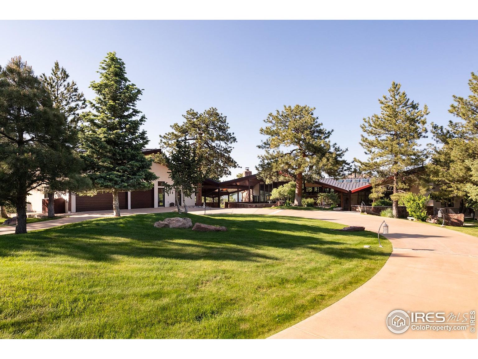 This is a front exterior view of a sprawling single-family home. The property features a well-manicured lawn, mature trees, and a long driveway leading to the house. The architecture includes a combination of modern and rustic elements, with a low-slung roofline and a mix of natural materials.