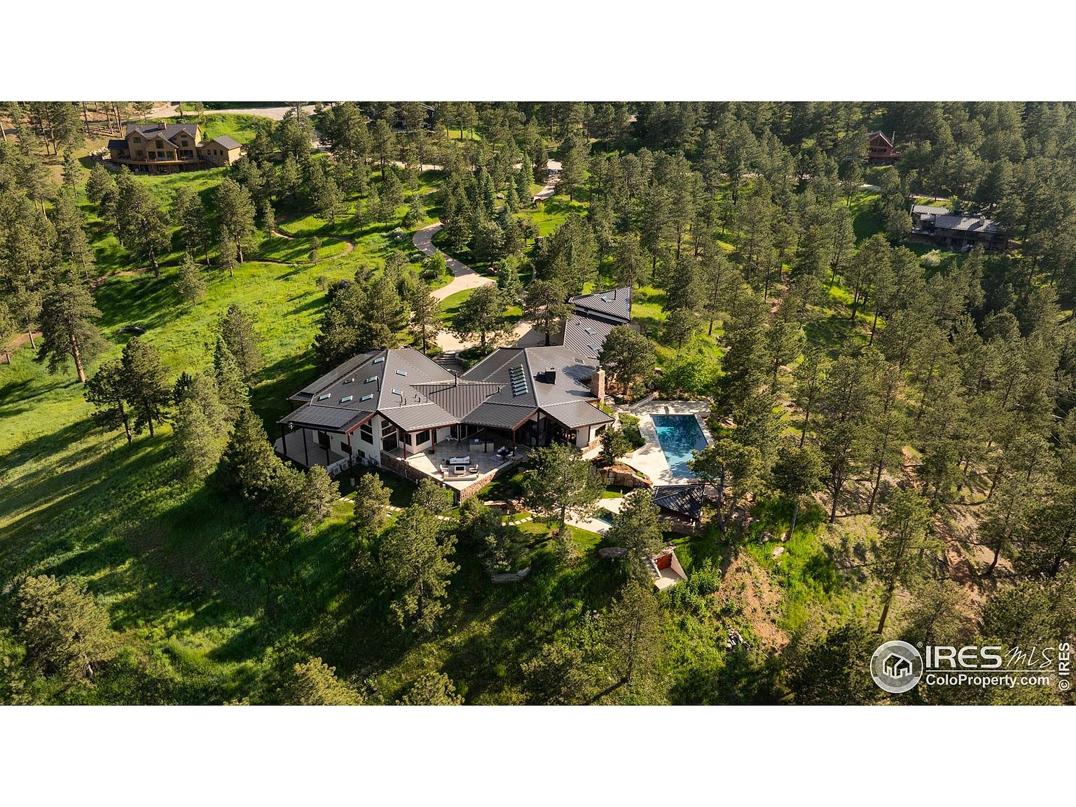 This aerial shot showcases a luxurious home nestled among lush greenery. The house features a modern architectural design with a dark roof and light-colored exterior walls. A swimming pool and patio area are visible, enhancing the property's appeal for outdoor living and entertainment.