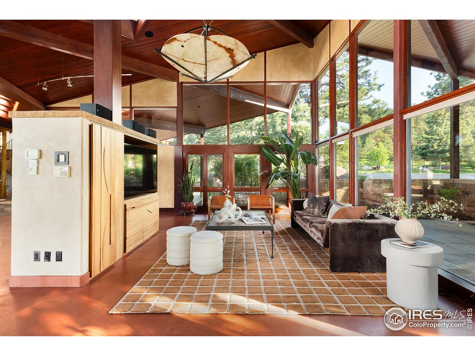 This is an interior shot of a living room featuring a high vaulted ceiling with wooden beams and large windows that offer an expansive view of the outdoors. The room is furnished with a brown sofa, white ottomans, a coffee table, and a side table, all arranged on a patterned rug. The style is modern with natural elements, creating a bright and airy atmosphere.