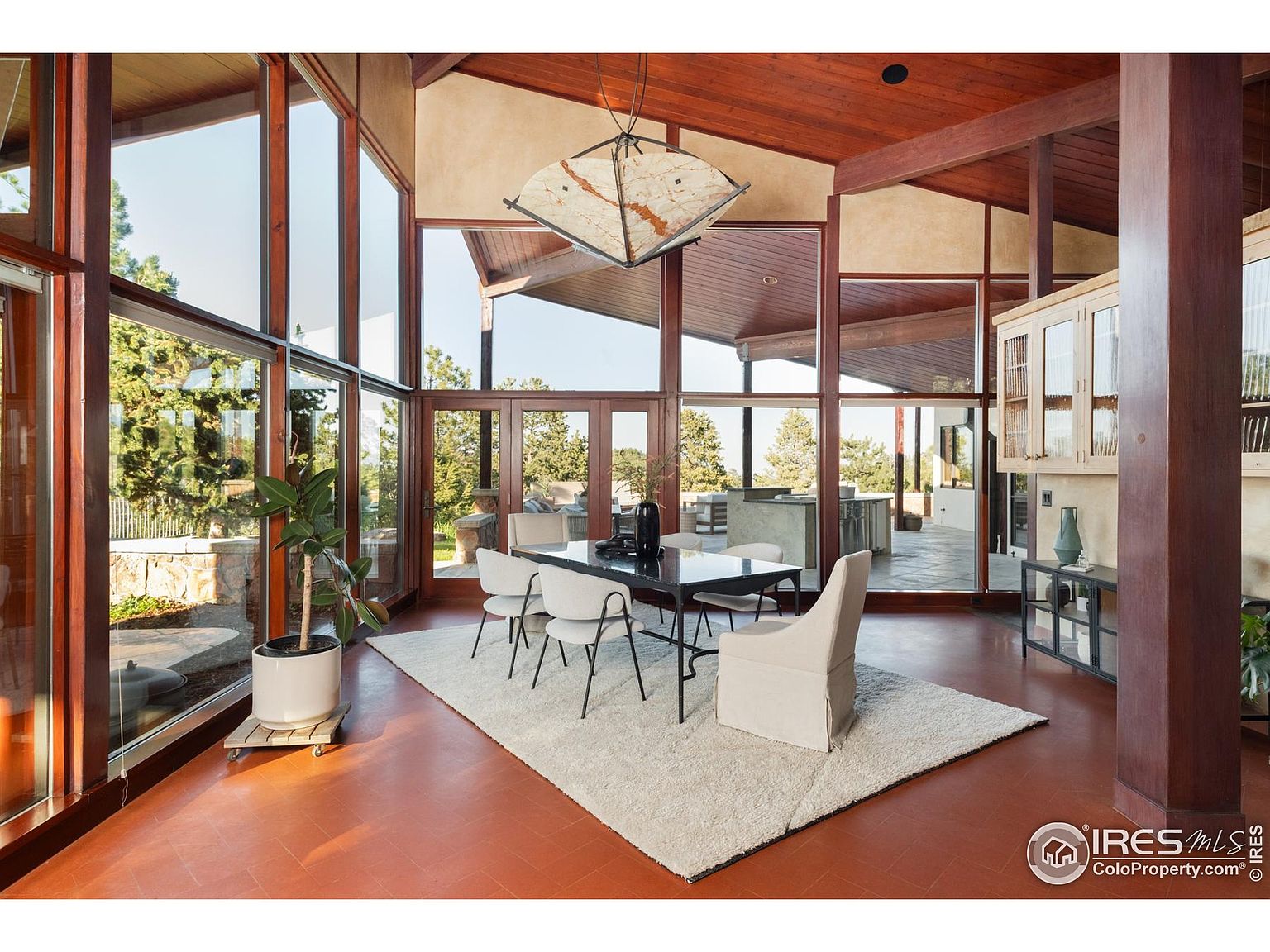This interior shot showcases a dining room with floor-to-ceiling windows offering an expansive view of the outdoors. The room features a dark dining table surrounded by modern chairs, set upon a light-colored rug. The architectural design includes exposed wooden beams and a unique chandelier, contributing to a sophisticated and airy atmosphere.