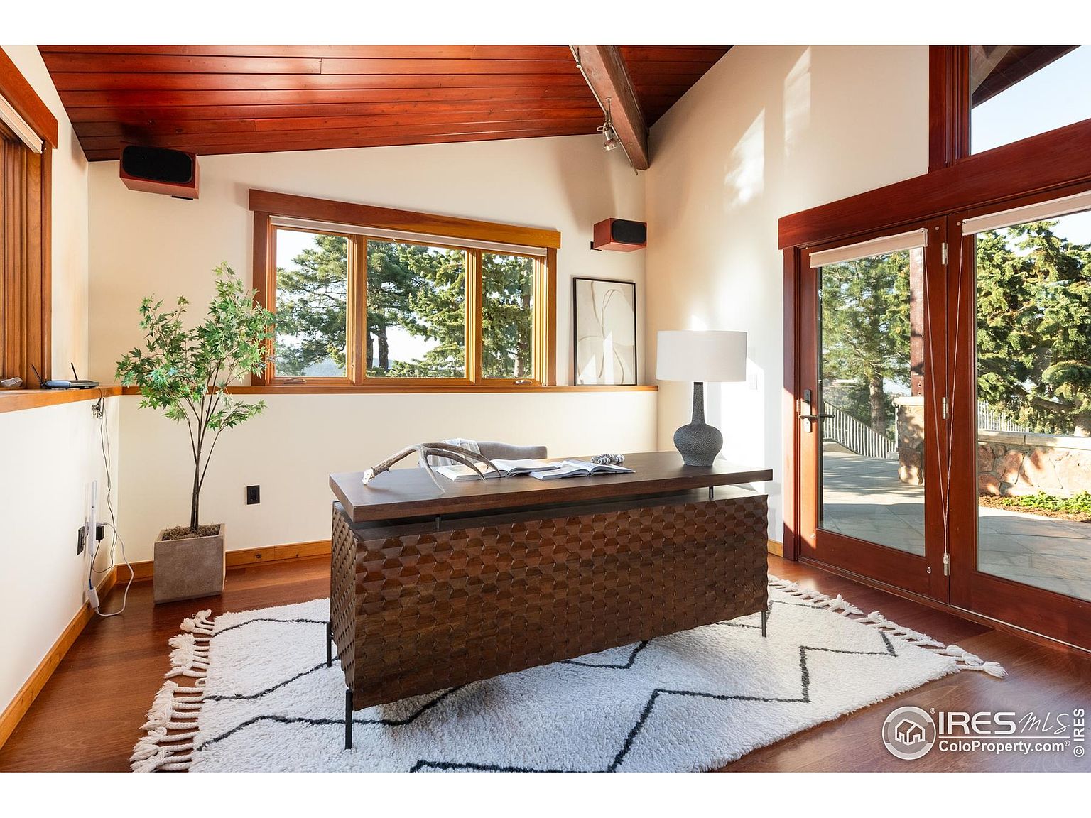 This is an interior shot of a home office featuring a unique wooden desk with a textured front, set on a white rug with a geometric pattern. The room has a warm, inviting feel with natural light streaming in through a large window and glass doors, showcasing the outdoor greenery. The ceiling is wood paneled, adding to the room's character.