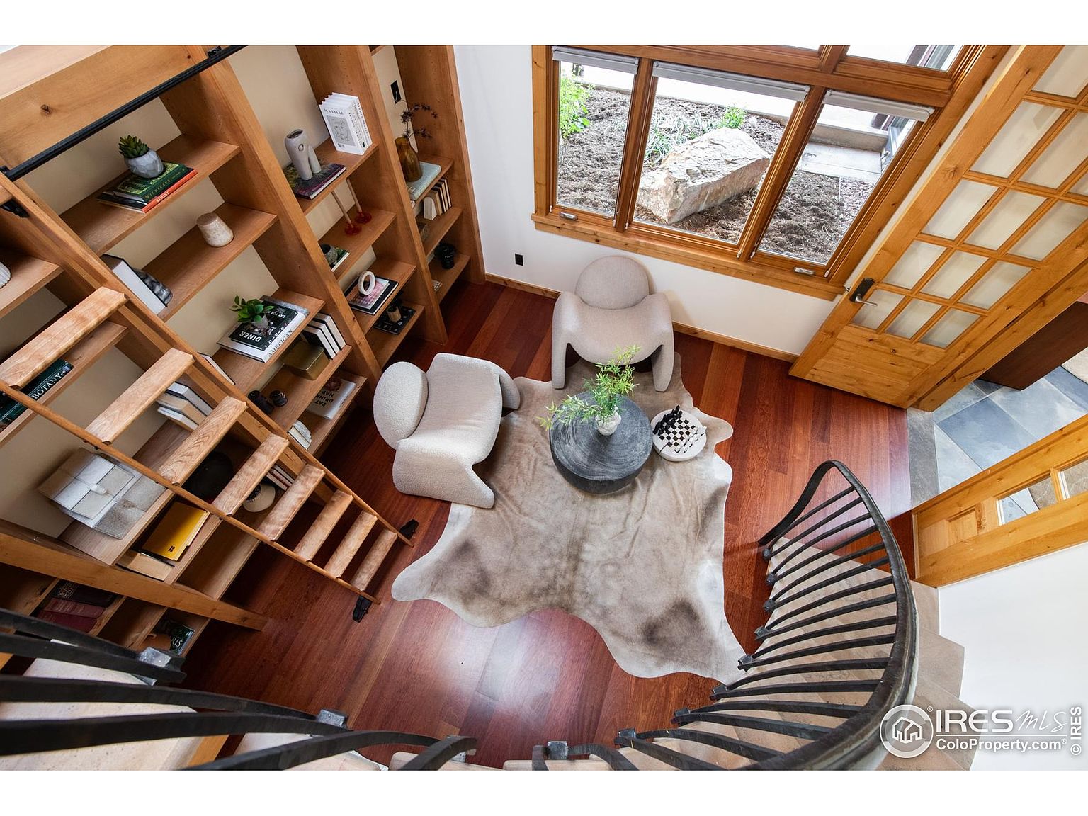 This image showcases an interior view of a hallway and staircase area. The space features hardwood flooring, a custom-built wooden bookshelf with a rolling ladder, and a unique cowhide rug. Two modern chairs and a round table with decorative items create a cozy seating area near a window, while a curved staircase with metal railings adds architectural interest.