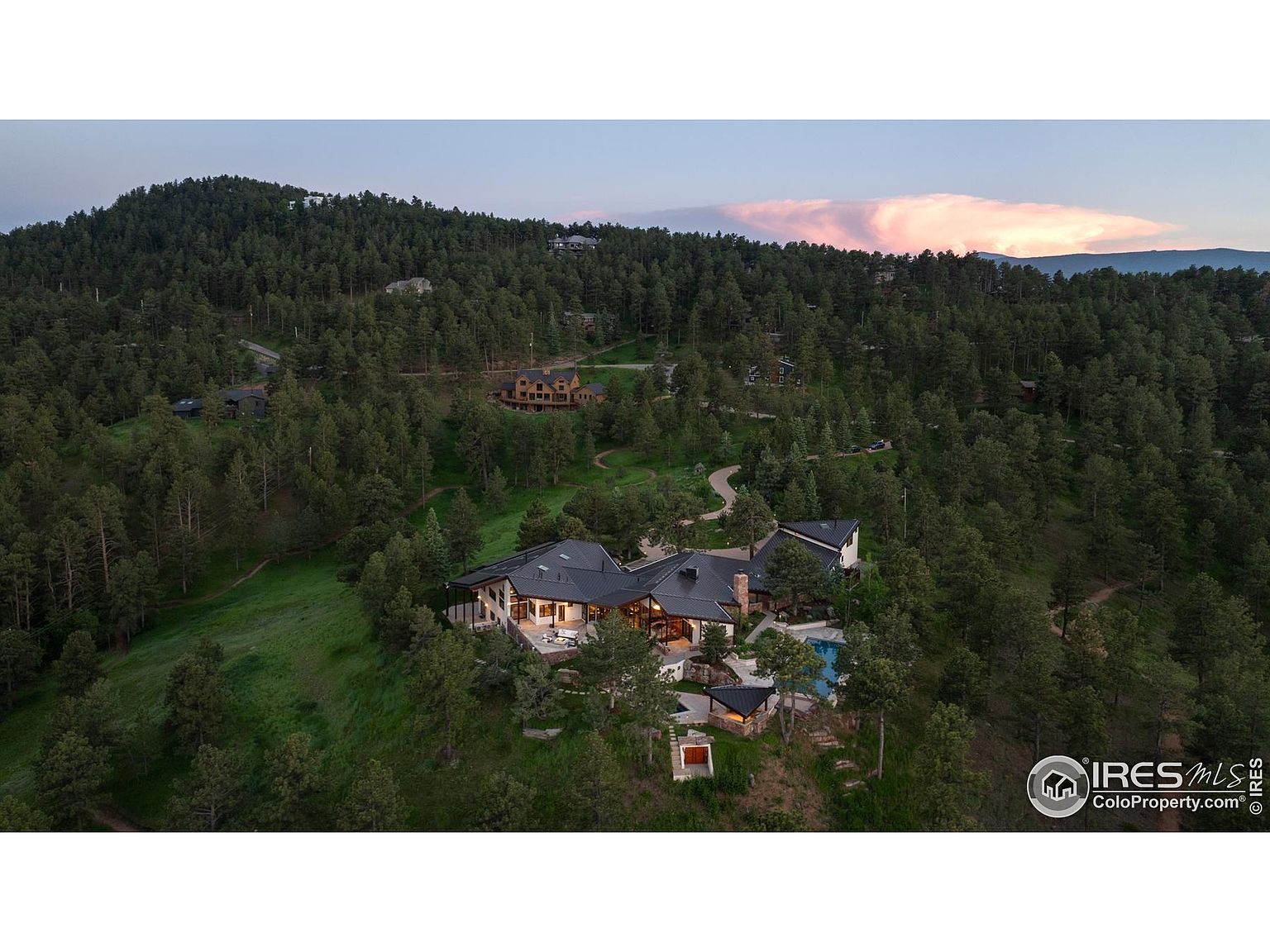 This aerial view showcases a luxurious home nestled among lush green trees on a hillside. The house features a dark roof, multiple outdoor living spaces, and a swimming pool. The surrounding landscape includes dense forest and a distant mountain range, creating a sense of privacy and natural beauty.