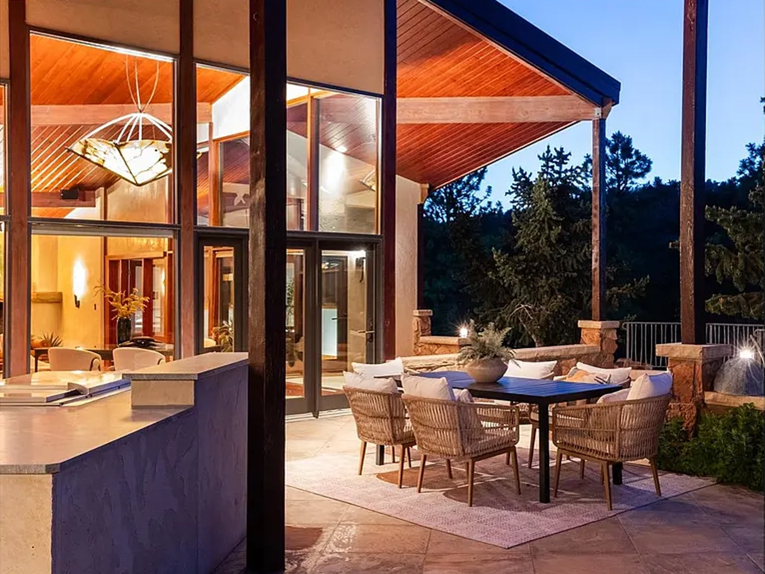 This image showcases an inviting covered patio area, perfect for outdoor dining and relaxation. The space features comfortable wicker chairs around a modern table, set on a stylish area rug. The patio seamlessly connects to the interior through large glass windows and doors, offering a blend of indoor and outdoor living with a natural backdrop of trees.