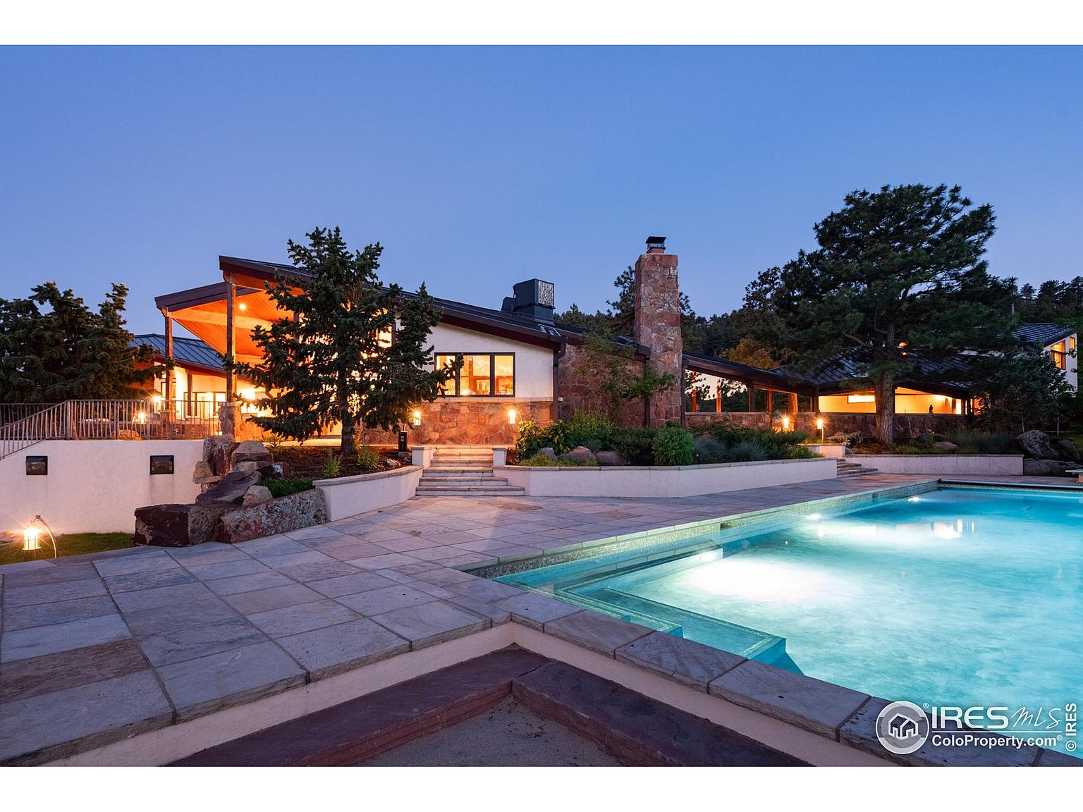 This image showcases a luxurious backyard featuring a large, inviting pool with clear blue water. The pool is surrounded by a stone patio, and the house is visible in the background, featuring a unique architectural design with a stone chimney and warm lighting. Mature trees and landscaping add to the serene and private atmosphere of the property.